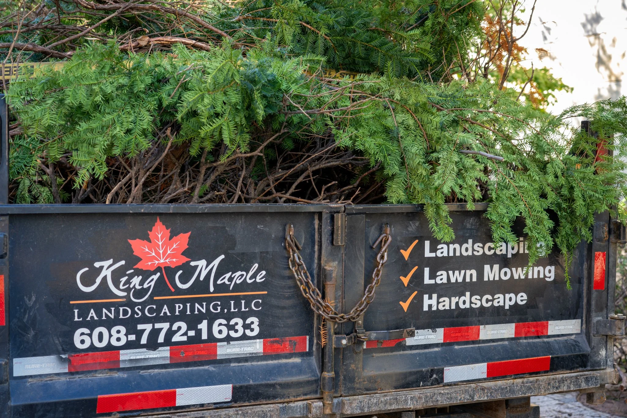 Trailer filled with freshly cut green tree branches and twigs, parked outdoors. The trailer has a sign advertising a landscaping business.