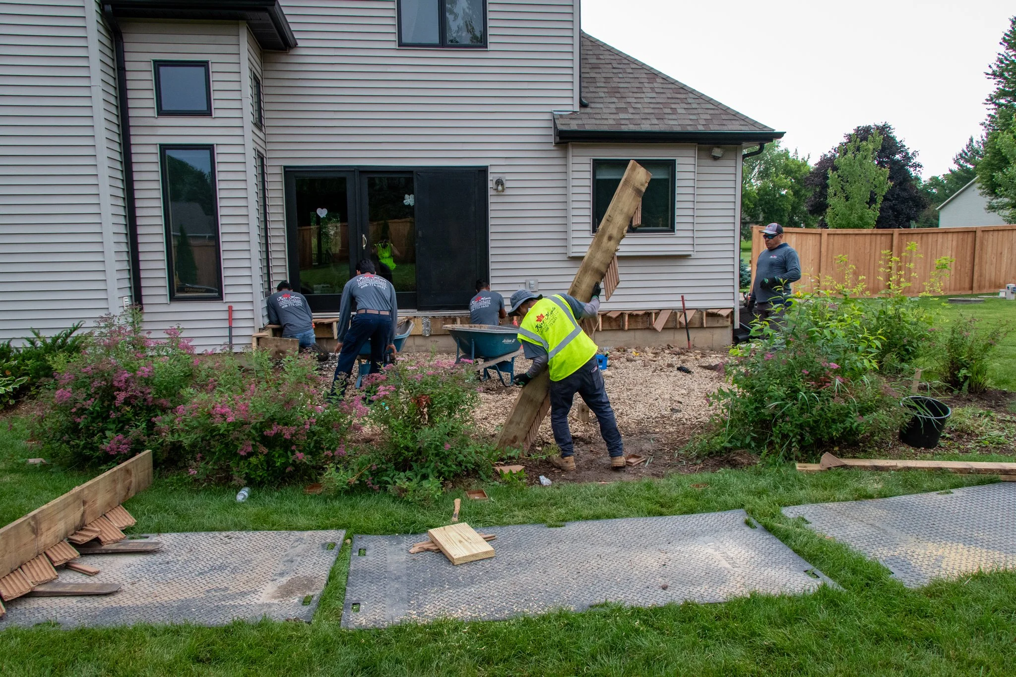 Workers demolishing or renovating a section of a house's backyard, with several people wearing high-visibility vests and work clothes, removing foundation and installing new supports, amidst plants and construction materials.
