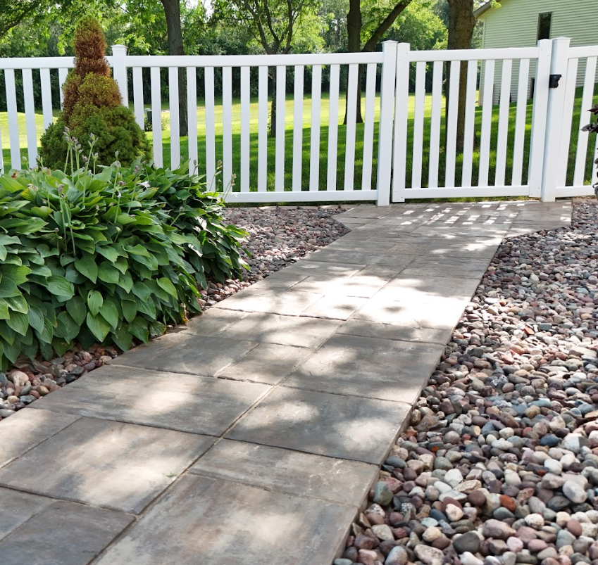 A paved walkway leading to a white fence gate, with green bushes and trees on the sides and a grassy yard in the background.