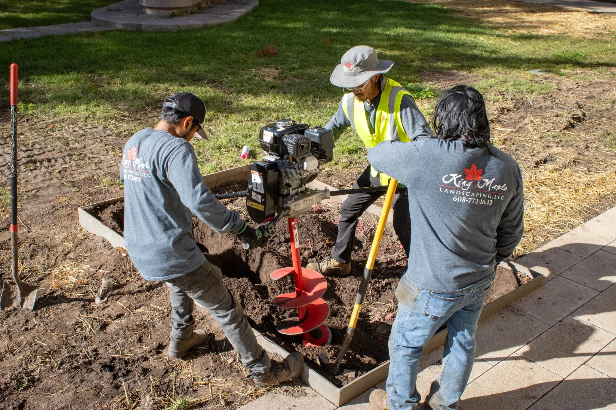 Three people working together planting or installing something in a garden bed, with a small motorized auger drill. They are wearing work clothes, and one is wearing a yellow vest and a wide-brim hat. There is grass and a paved walkway in the backgro
