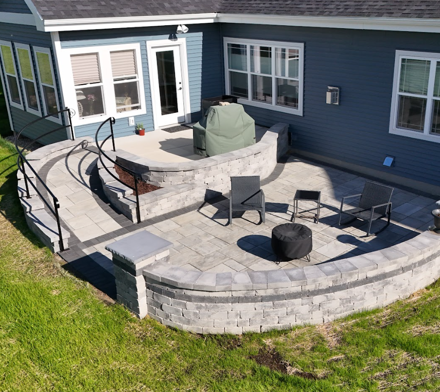 A backyard patio with a curved stone wall, multiple chairs, a small table, and a covered grill, attached to a blue house with large windows.