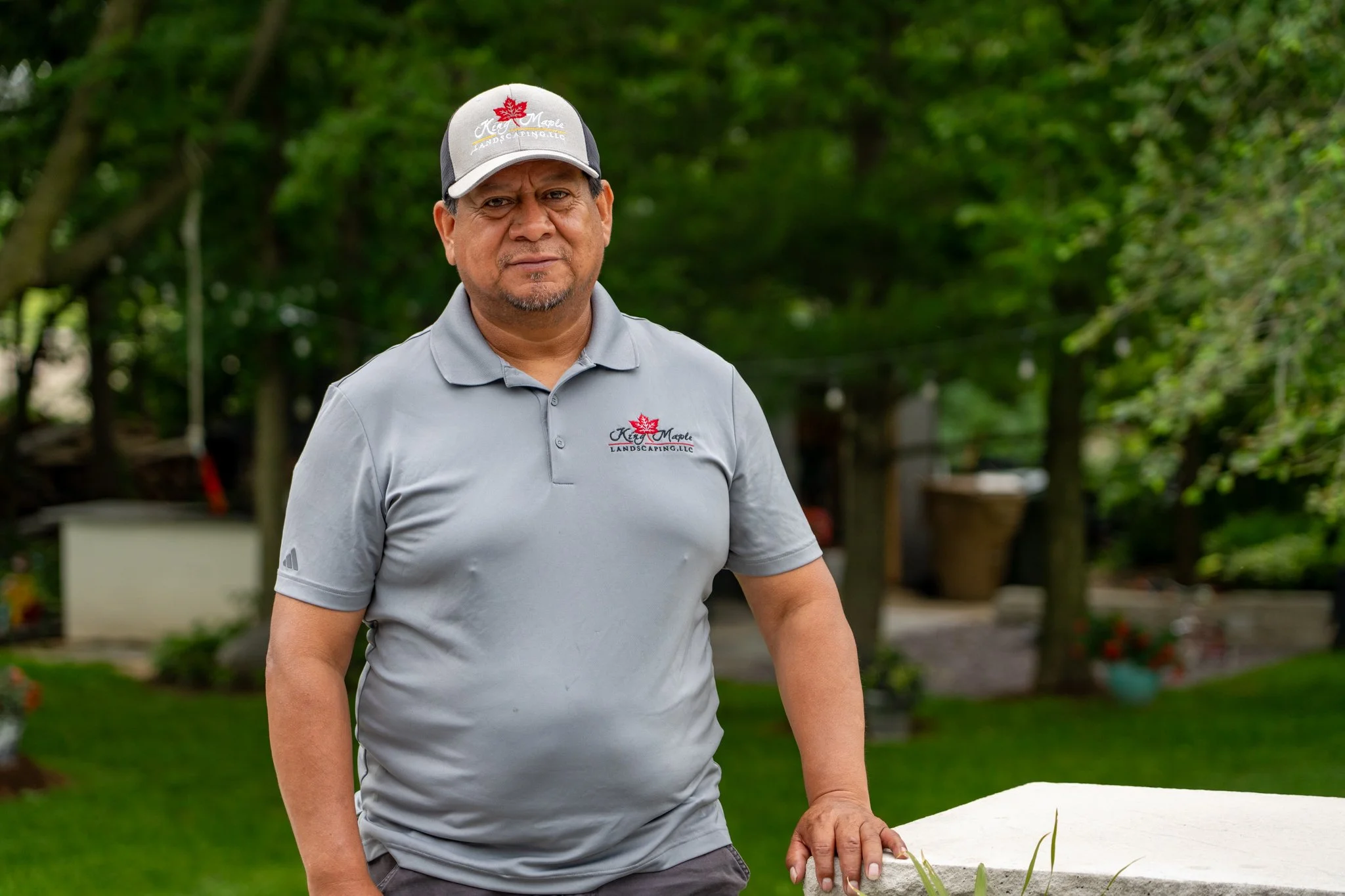 A man in a gray polo shirt and a baseball cap stands outdoors in a garden or park with green trees in the background.