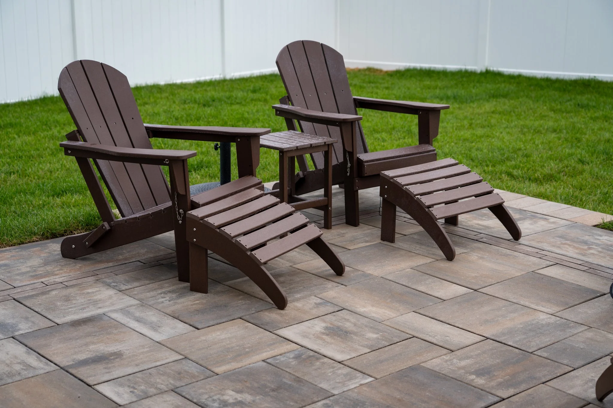 Two brown wooden Adirondack chairs and a small matching side table on a stone patio, with green grass and a white fence in the background.