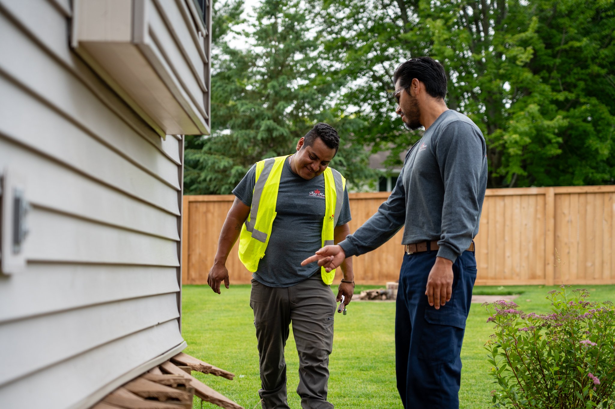 Two King Maple employees discussing outside near a beige house with horizontal siding, surrounded by green trees and a wooden fence, with purple flowers in the foreground.