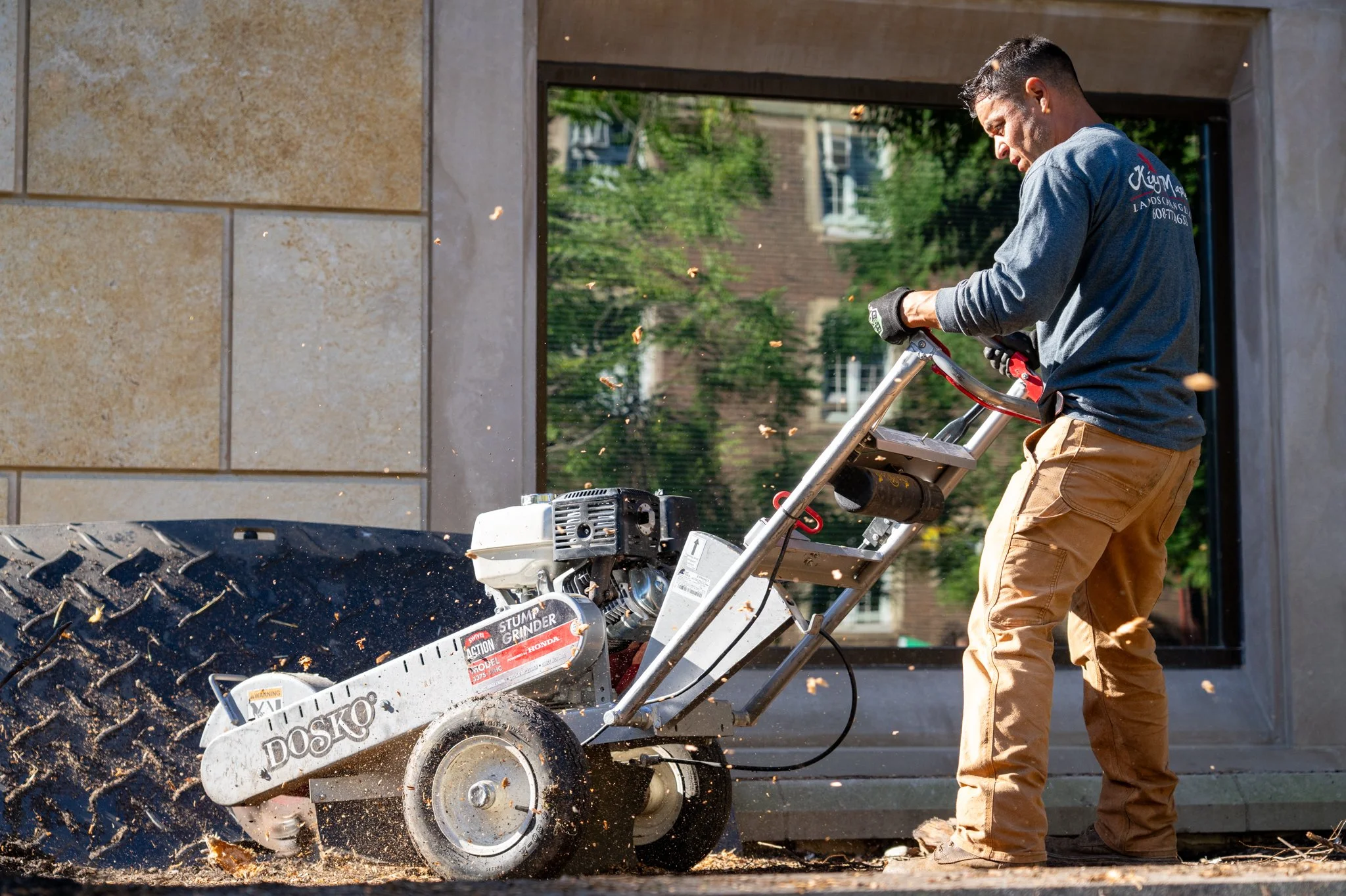 Man in gray long-sleeve shirt and tan work pants operating a large stump grinder on a sidewalk, with a building and greenery in the background.