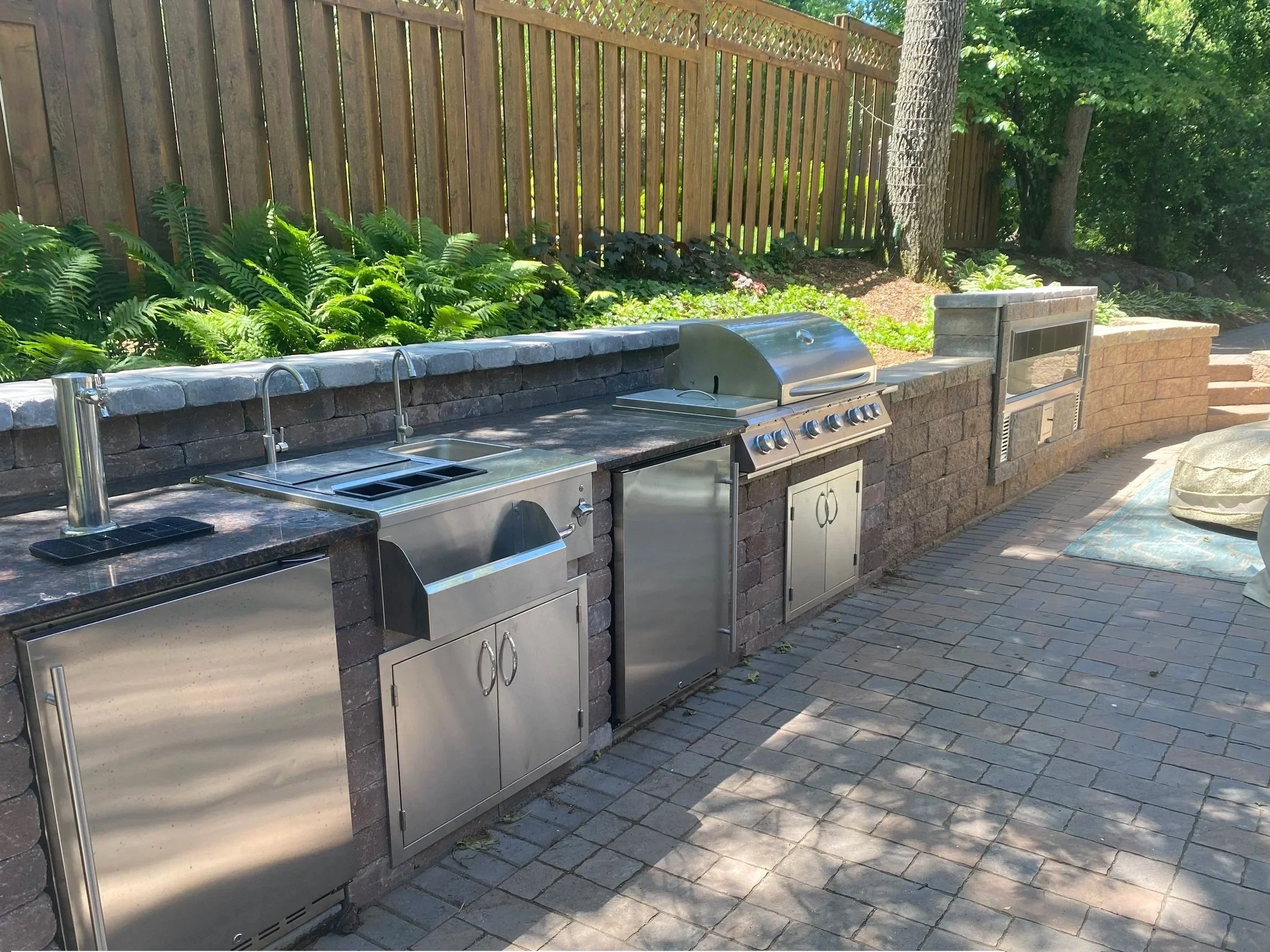 Outdoor kitchen with stainless steel appliances, including a grill and sink, built into a brick counter. There is a wooden fence and trees in the background, with lush green plants along the fence.