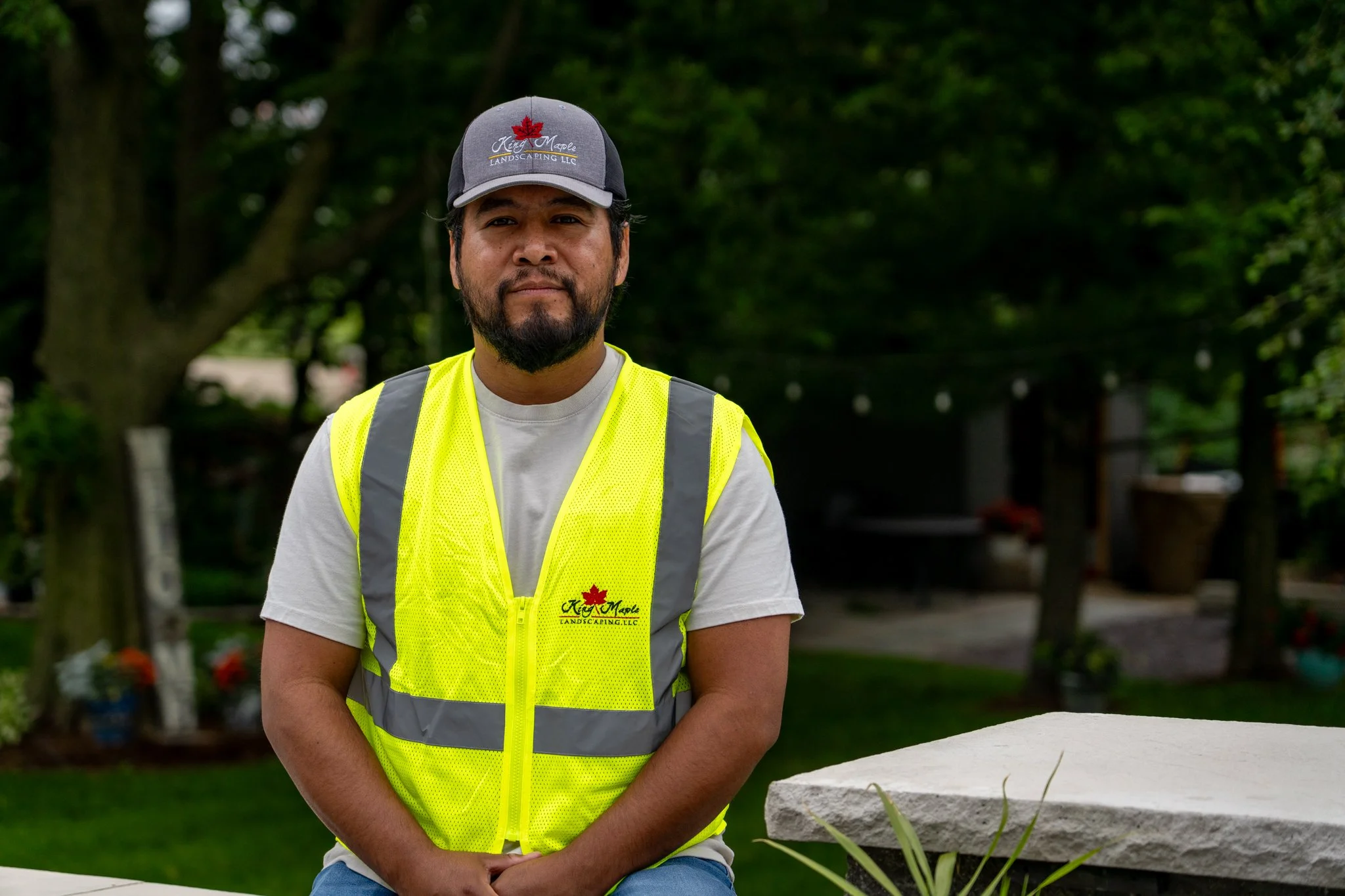 King Maple employee wearing a King Maple hat and a yellow safety vest sitting outdoors with trees and greenery in the background.