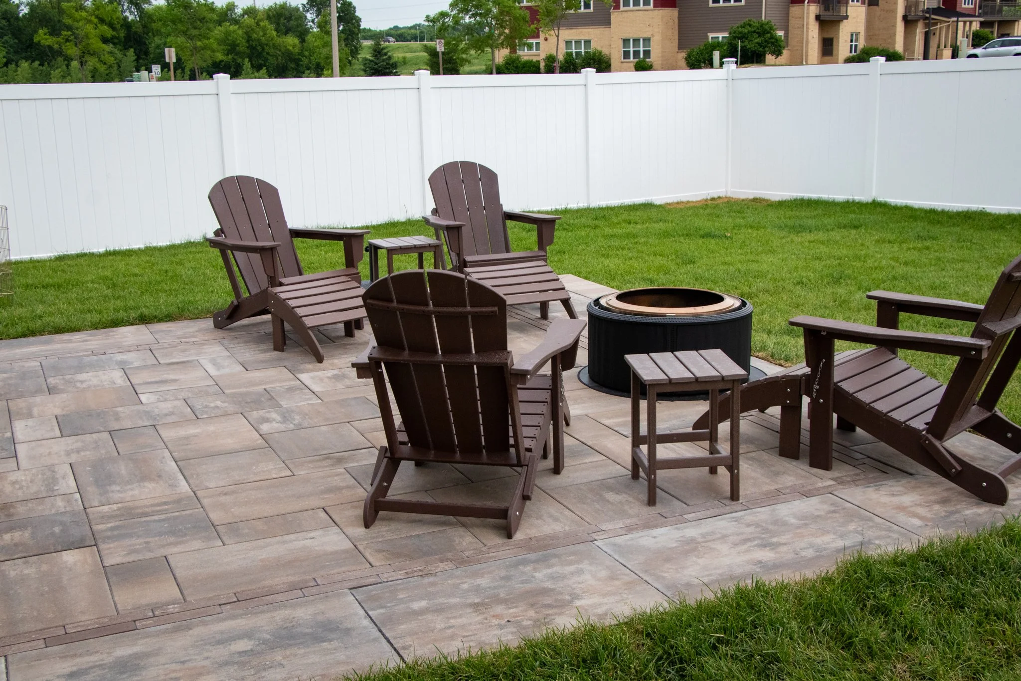 Backyard with a patio featuring six wooden Adirondack chairs arranged in a circle around a fire pit. The yard is enclosed with a white vinyl privacy fence, and there is green grass and a row of trees beyond the fence. In the background, apartment bui