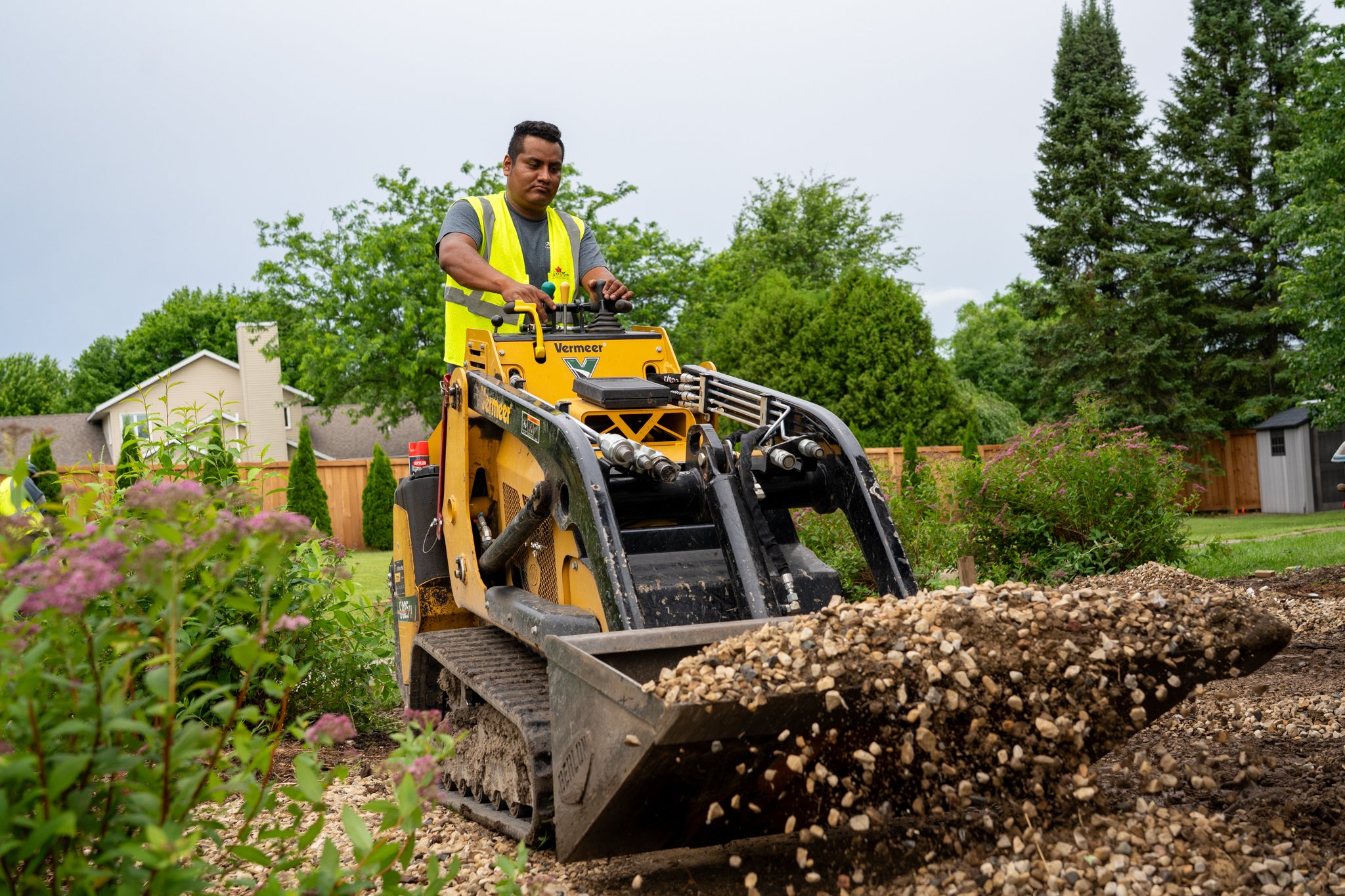 King Maple Employee operating a small yellow construction vehicle with the brand name 'Vermeer' in a backyard garden. The vehicle is pushing a load of small rocks and gravel. The background features green trees, a wooden fence, and a house.