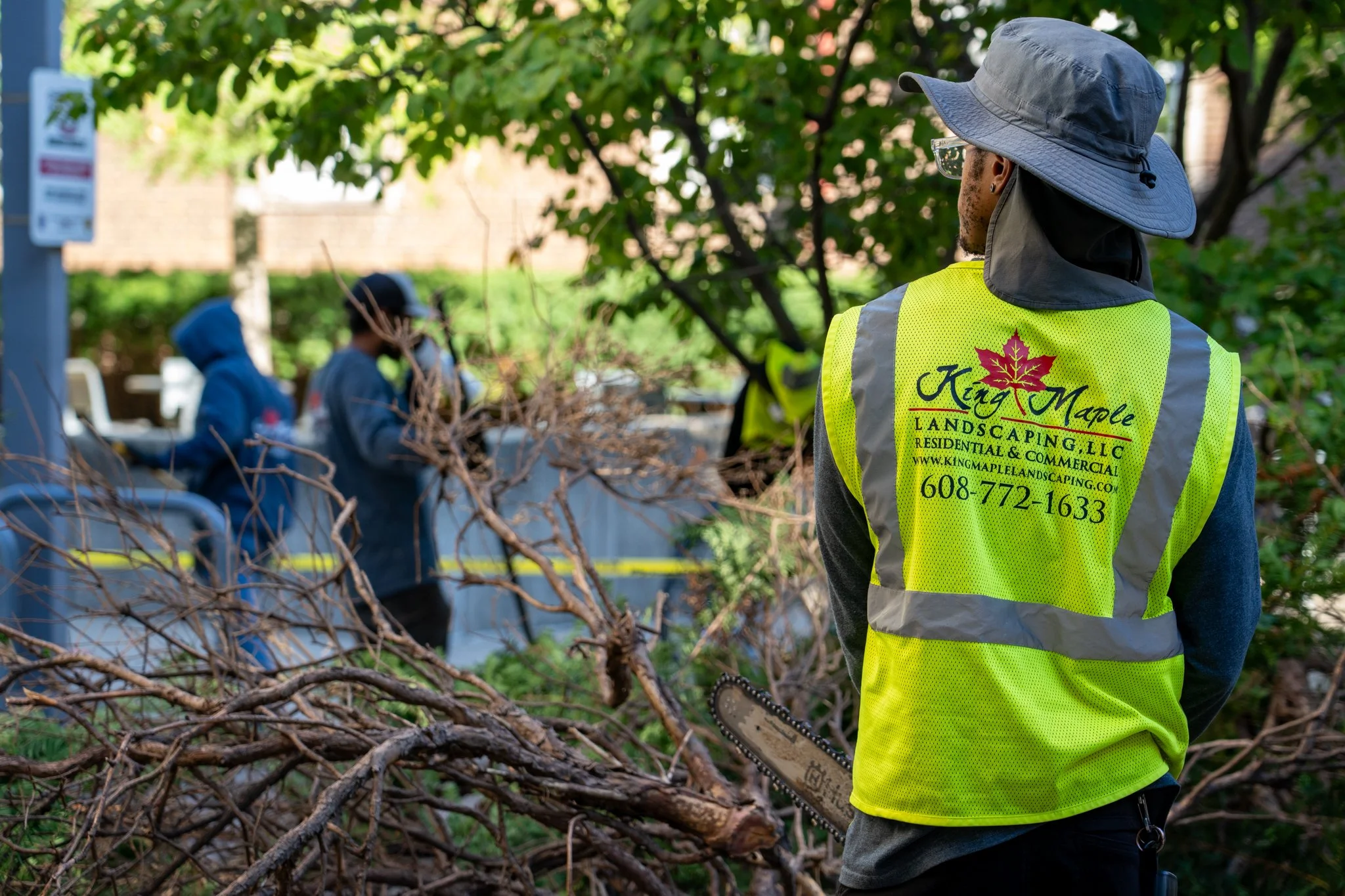 A King Maple Employee in a yellow King Maple branded safety vest and wide-brimmed hat using a chainsaw to cut branches in a yard, with two other people in hoodies in the background.