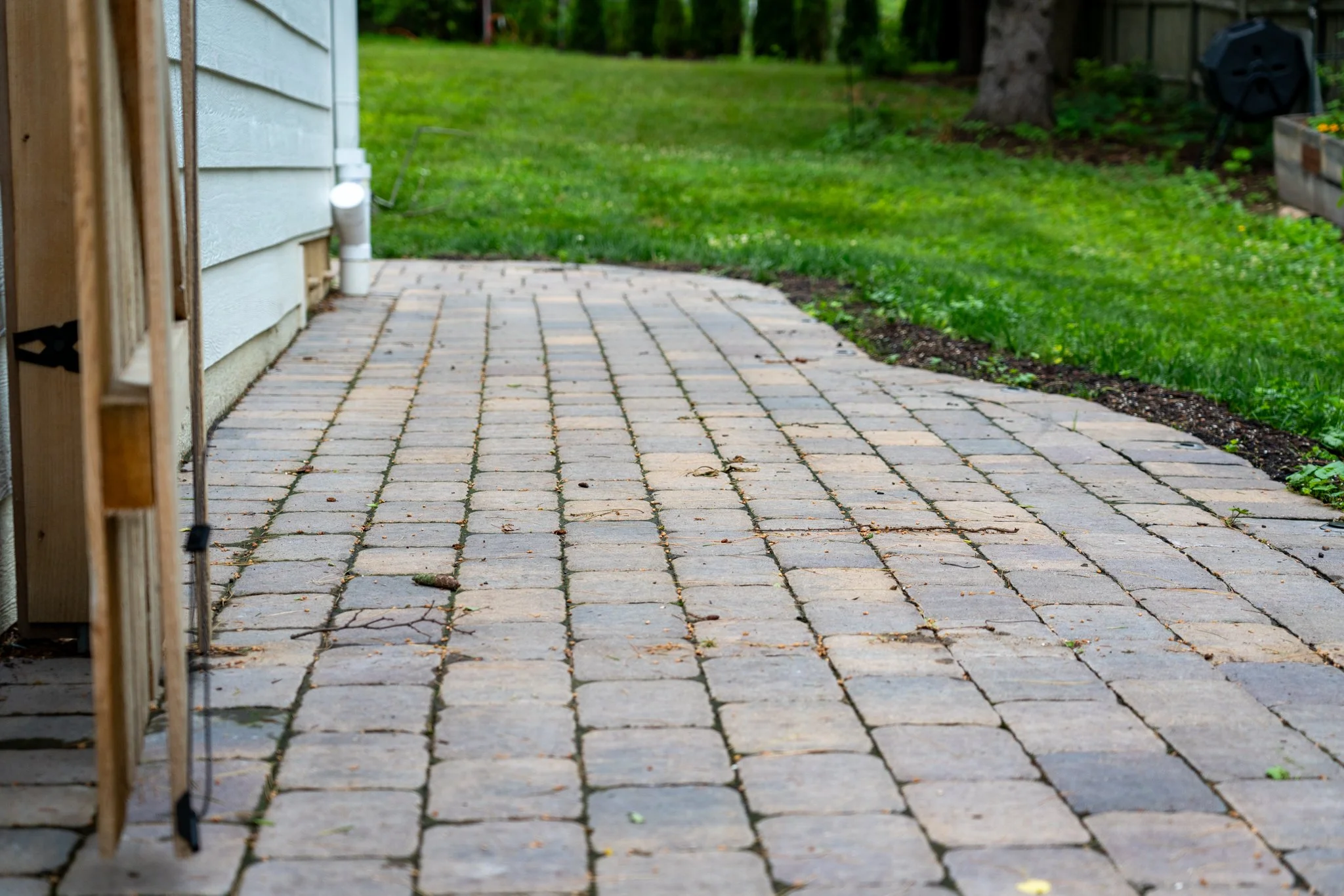 Backyard patio with stone pavers, white house siding on the left, green grass and trees in the background.