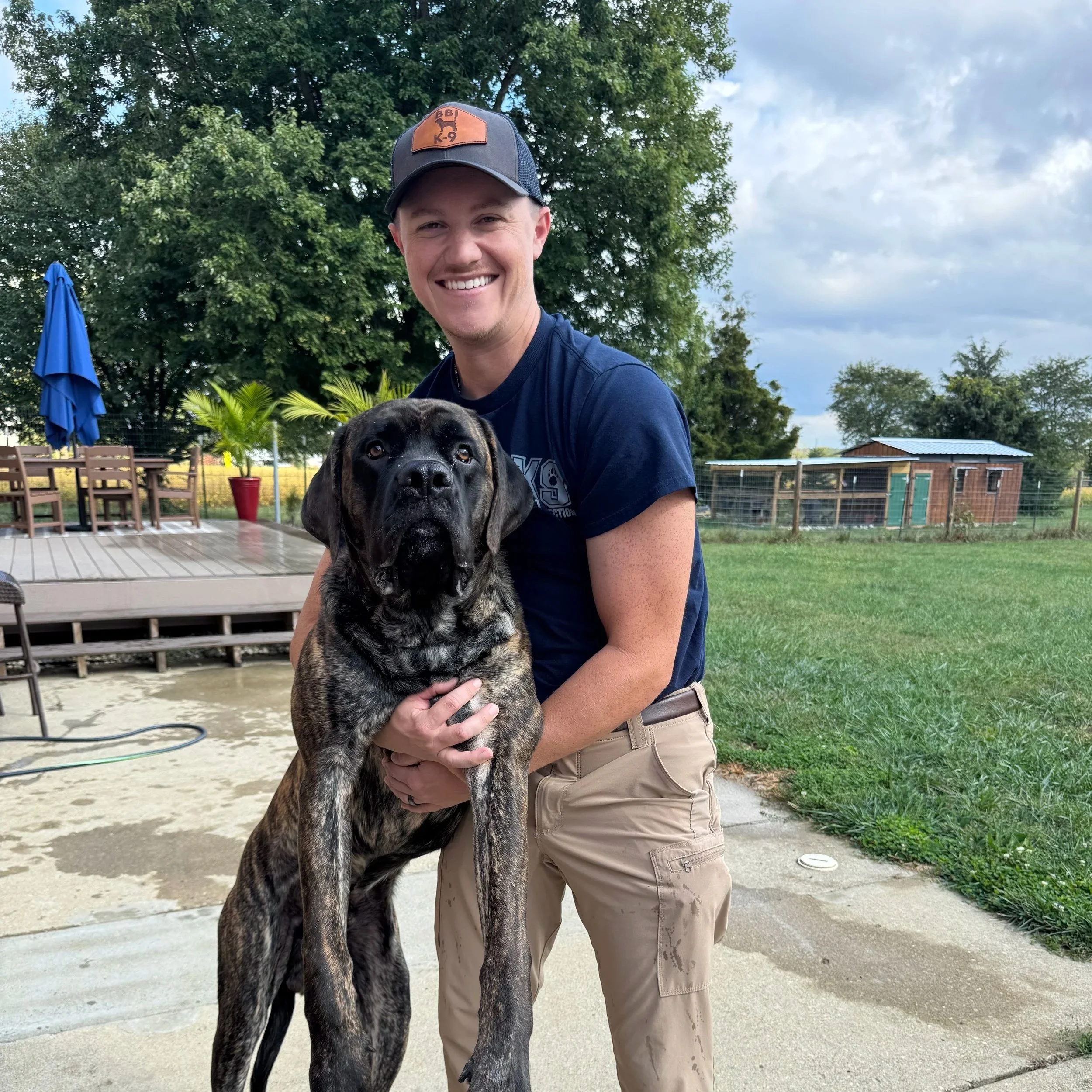 A smiling man in a navy shirt and tan shorts holding a large brindle-coated dog outdoors, with trees, a blue patio umbrella, and a fenced yard in the background.