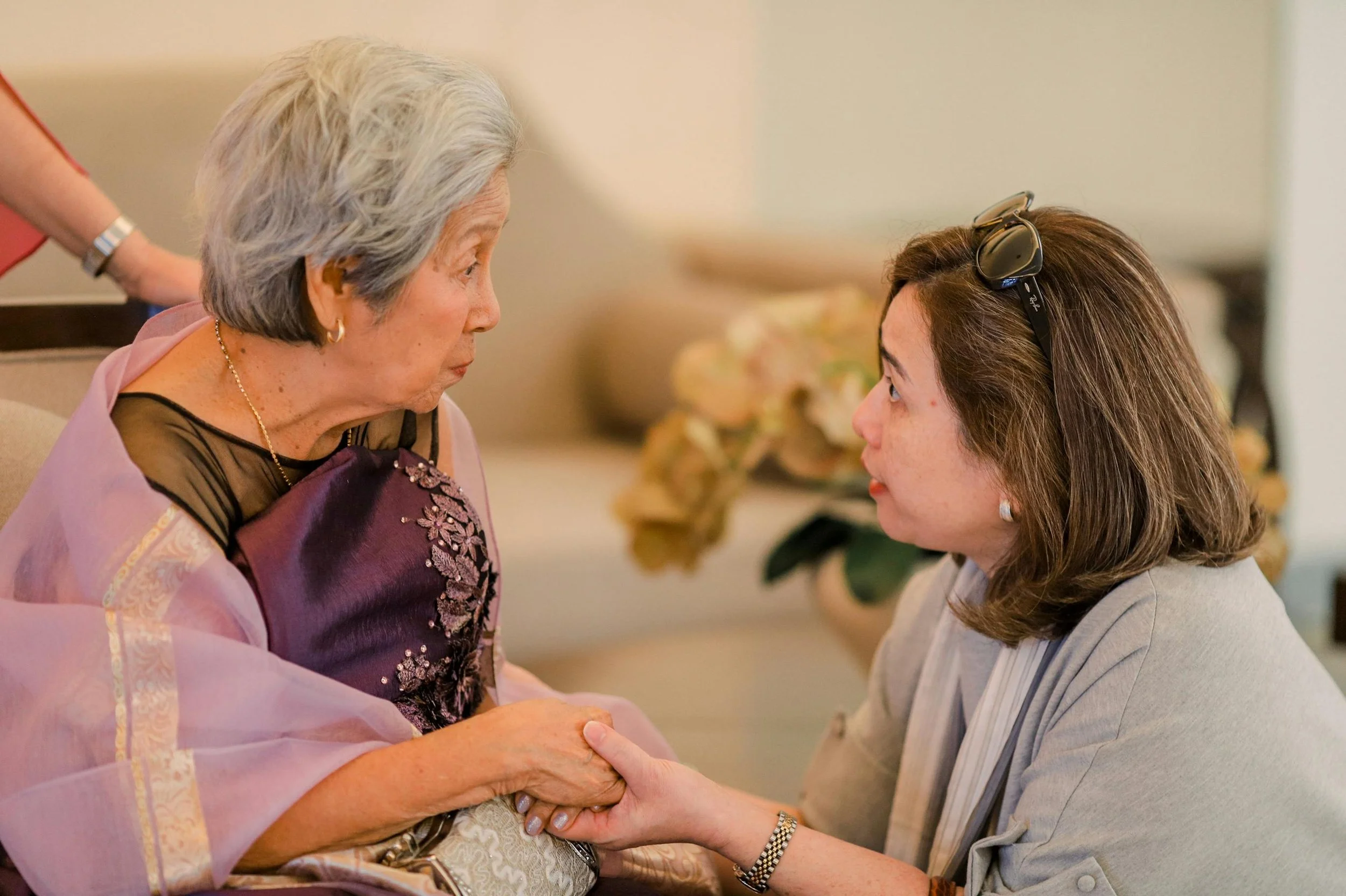 An elderly woman and a younger woman are holding hands and looking into each other's eyes in a warm, intimate setting.