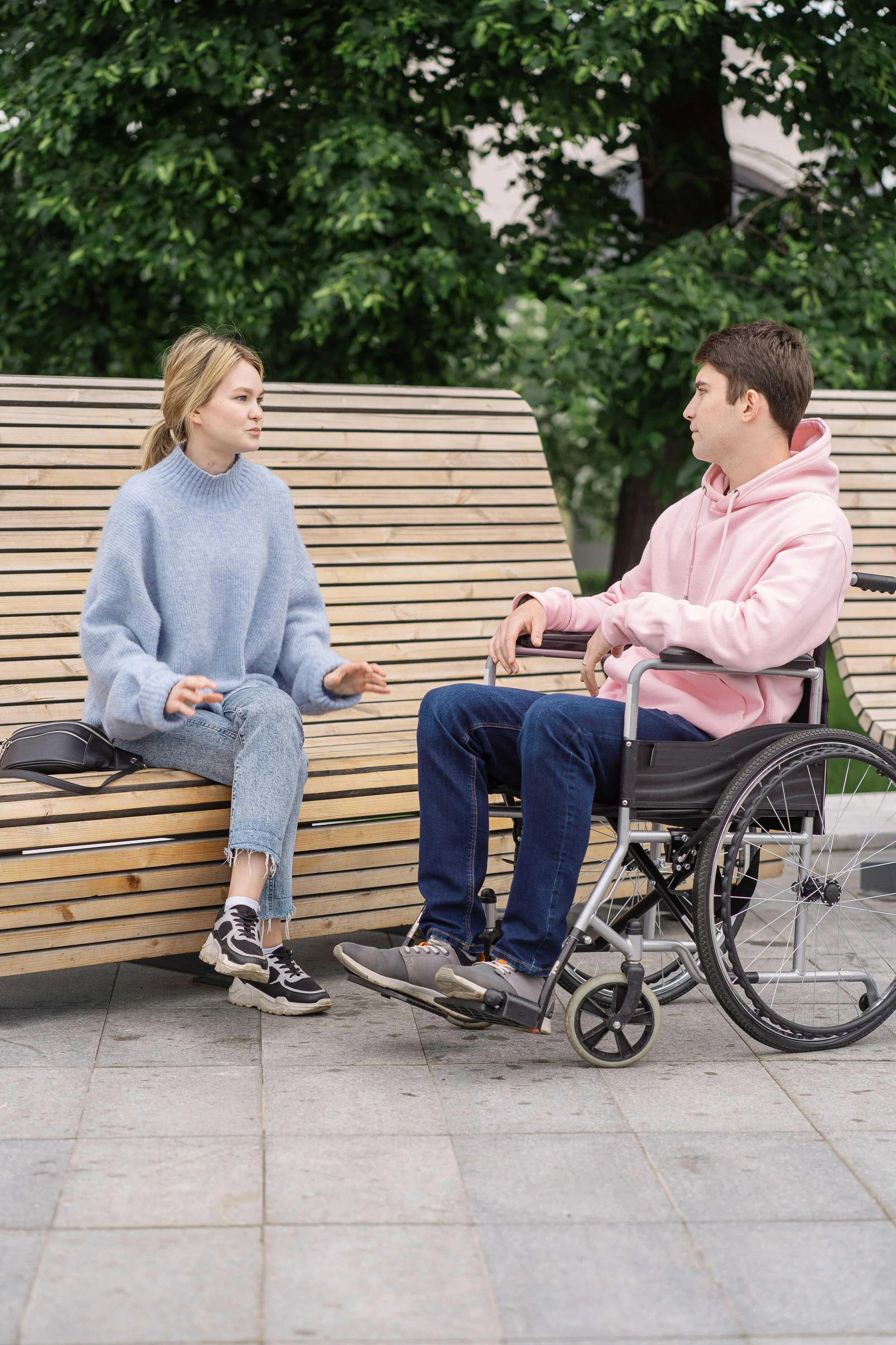 A young woman and a young man in a wheelchair have a conversation on a park bench, surrounded by green trees.