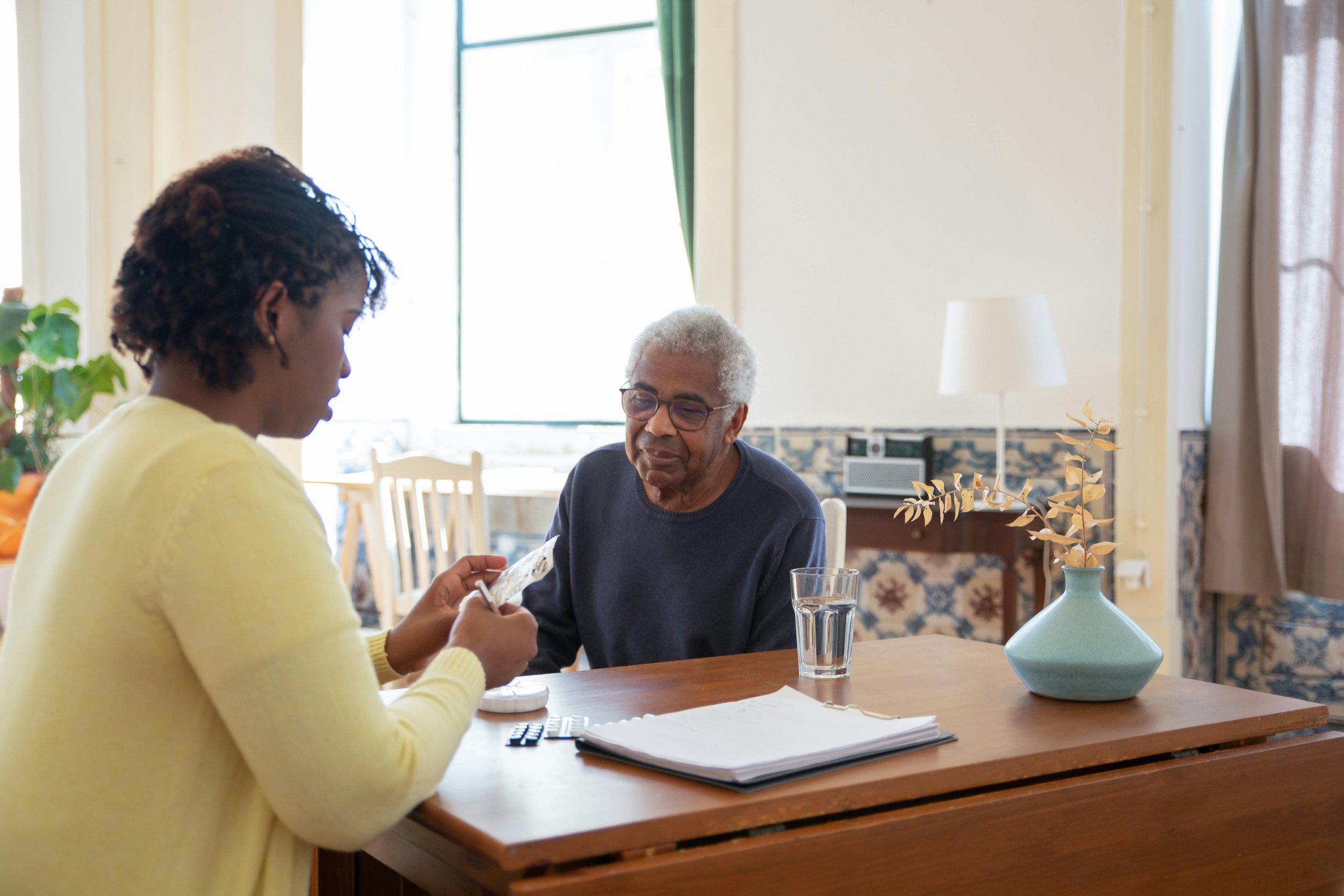 A woman in a yellow sweater showing money to an elderly man with gray hair and glasses, sitting at a wooden table in a bright room.