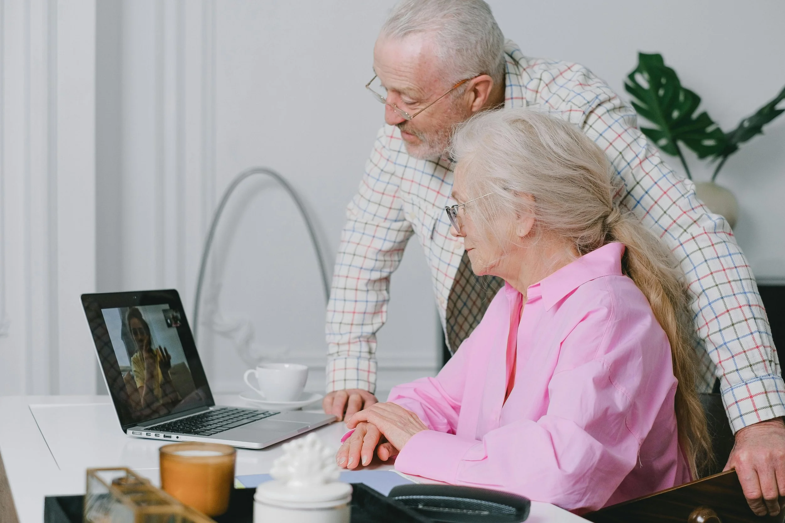 An elderly woman in pink shirt sitting at a table using a laptop, with an elderly man in a checkered shirt standing beside her, both looking at the laptop screen.