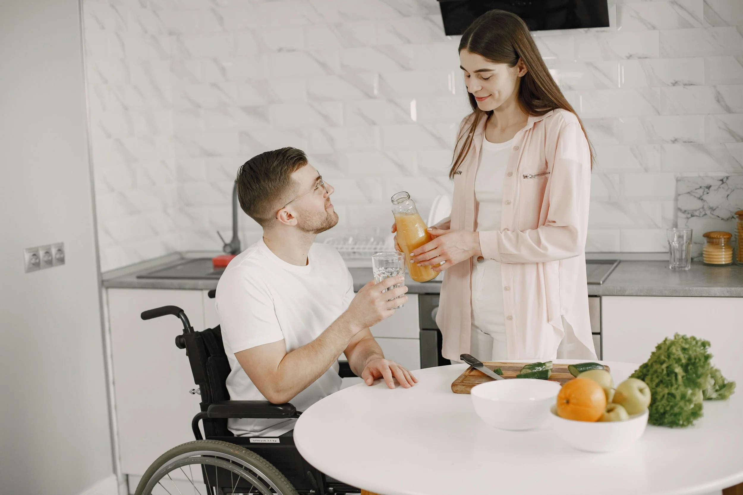 A woman is pouring orange juice into a glass held by a man in a wheelchair at a kitchen table. The table has a bowl of fruits and a knife on a cutting board.