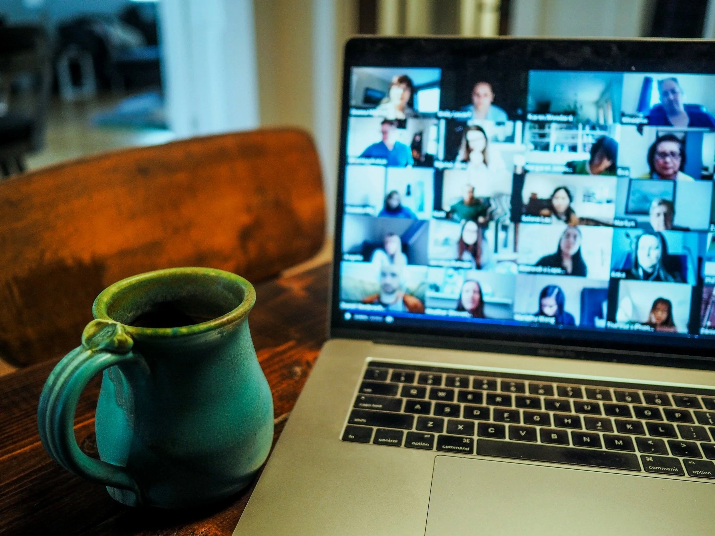 A ceramic mug with a handle sits on a wooden table, with a laptop displaying a video conference with multiple participants in the background.