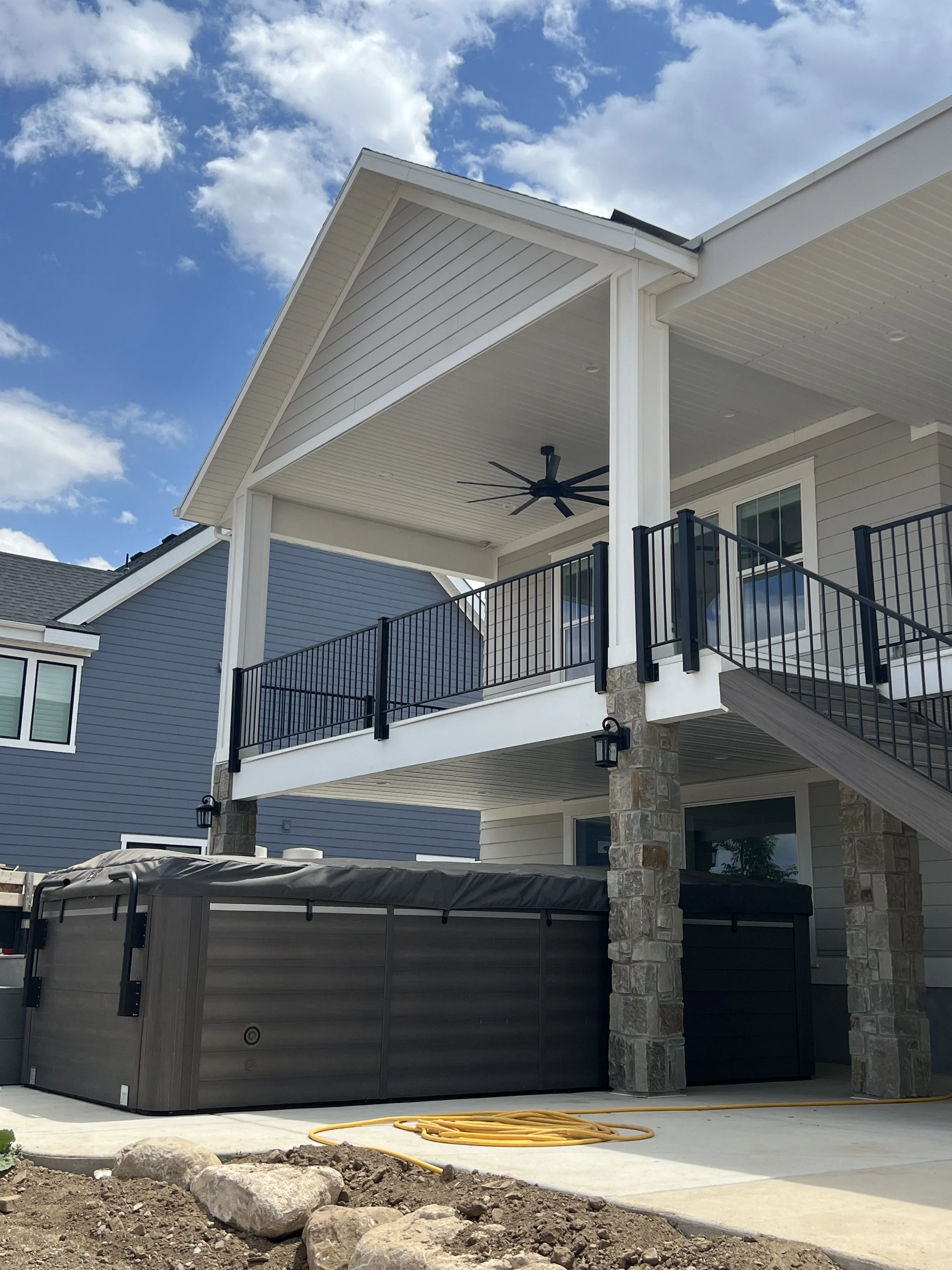 Backyard of a modern house with a covered deck, black railing, stone pillars, and a hot tub.