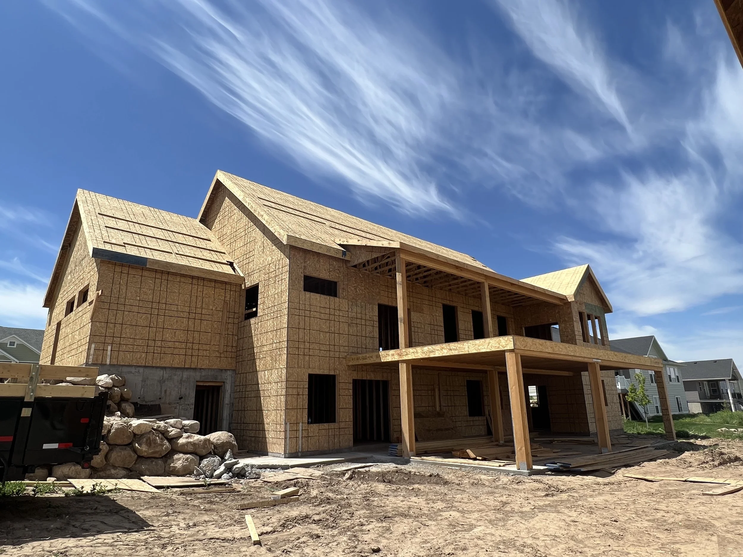 Under construction two-story house with wooden framing, multiple windows, and a porch, set against a clear blue sky with wispy clouds.