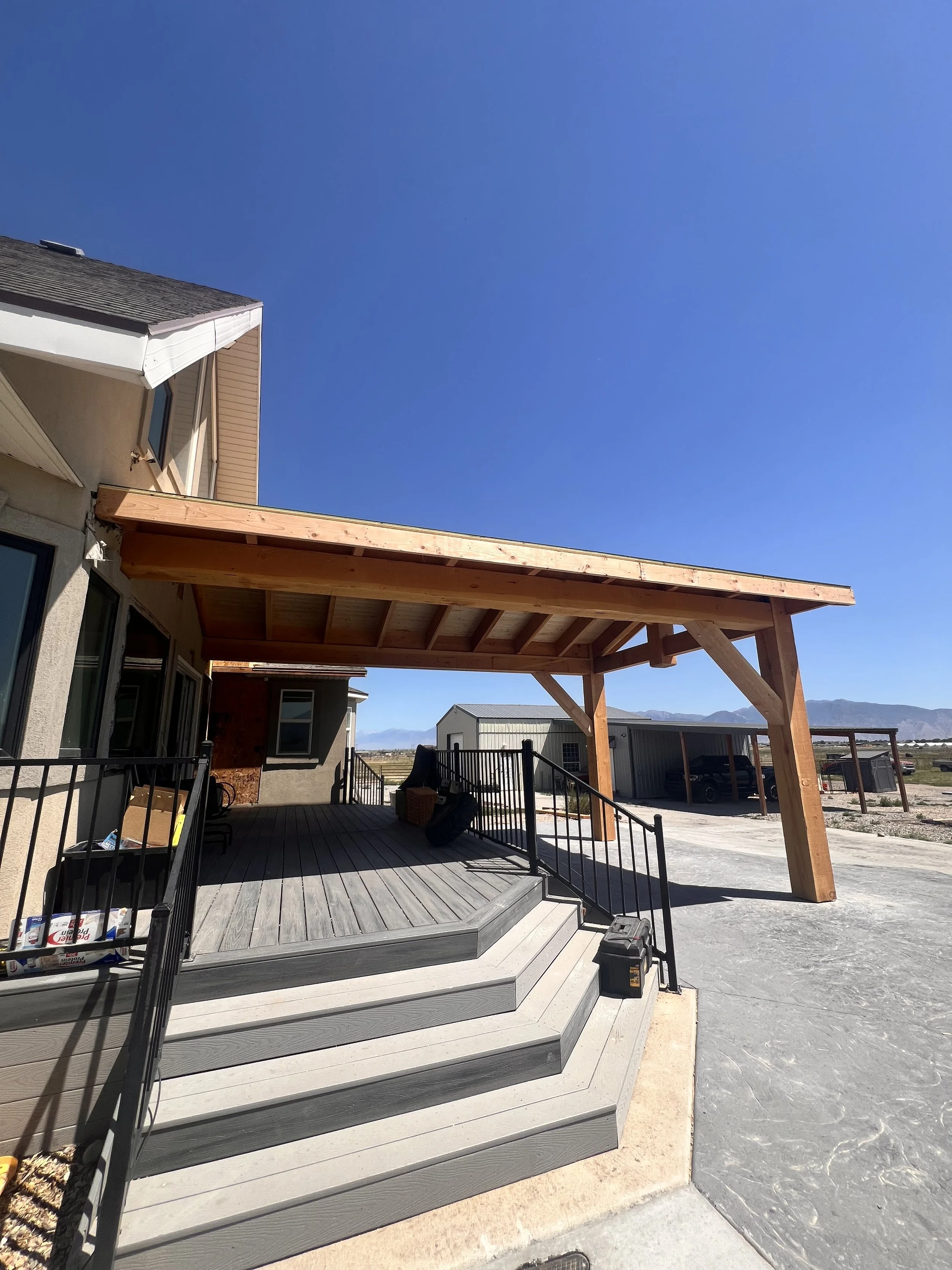 The image shows a residential outdoor area with a new wooden covered patio attached to a house, with stairs leading down to a concrete driveway. The sky is clear and blue.