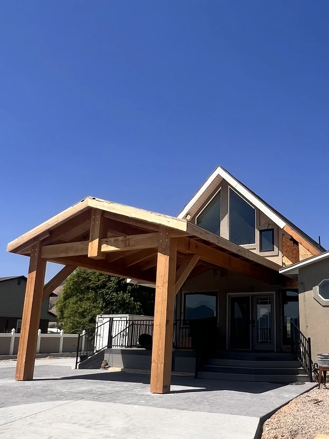 House with a wooden carport under construction, featuring supportive posts and a sloped roof, set against a clear blue sky.