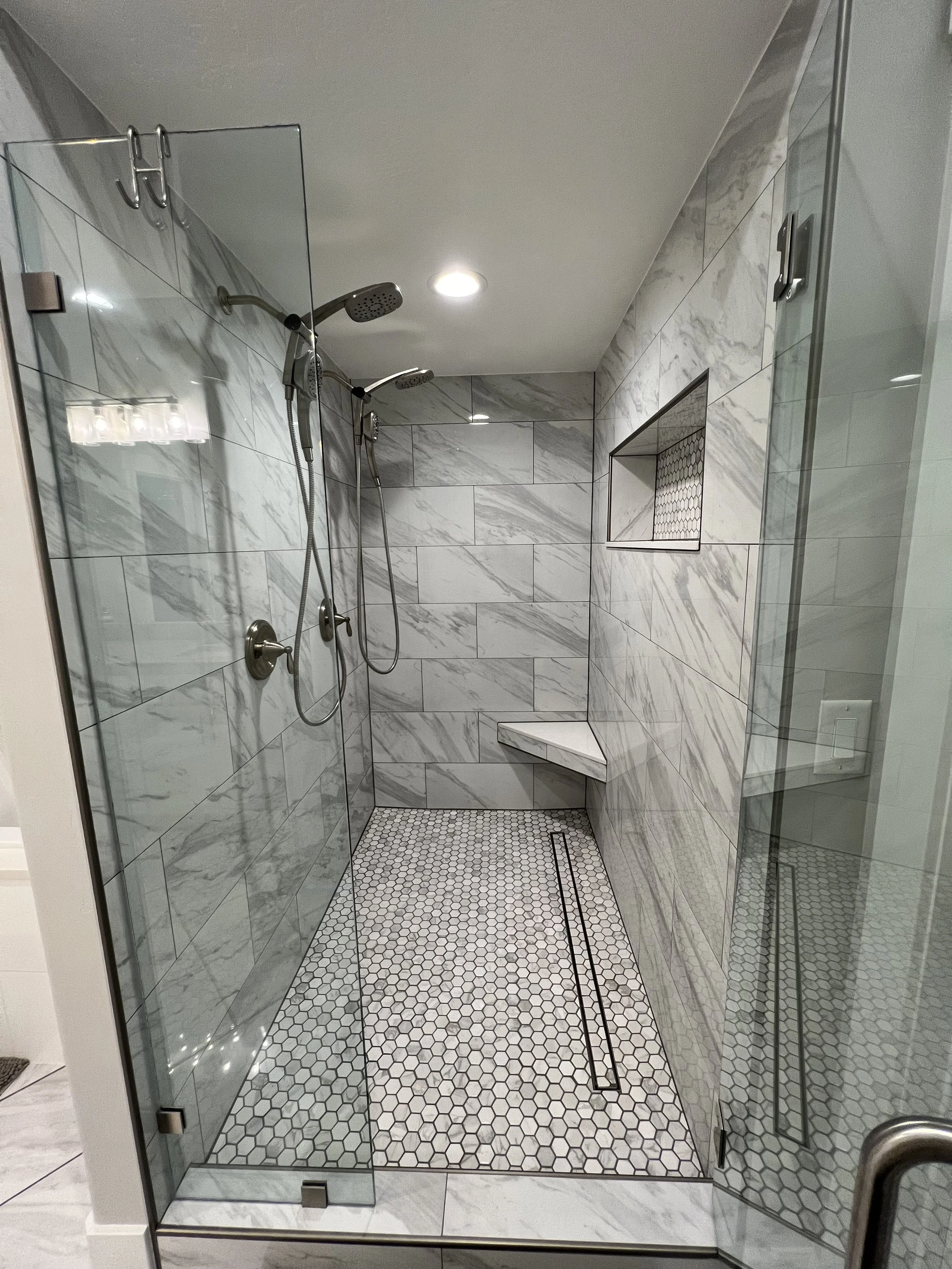 Modern walk-in shower with marble tiles, two showerheads, a built-in niche, and a bench, enclosed with glass doors.