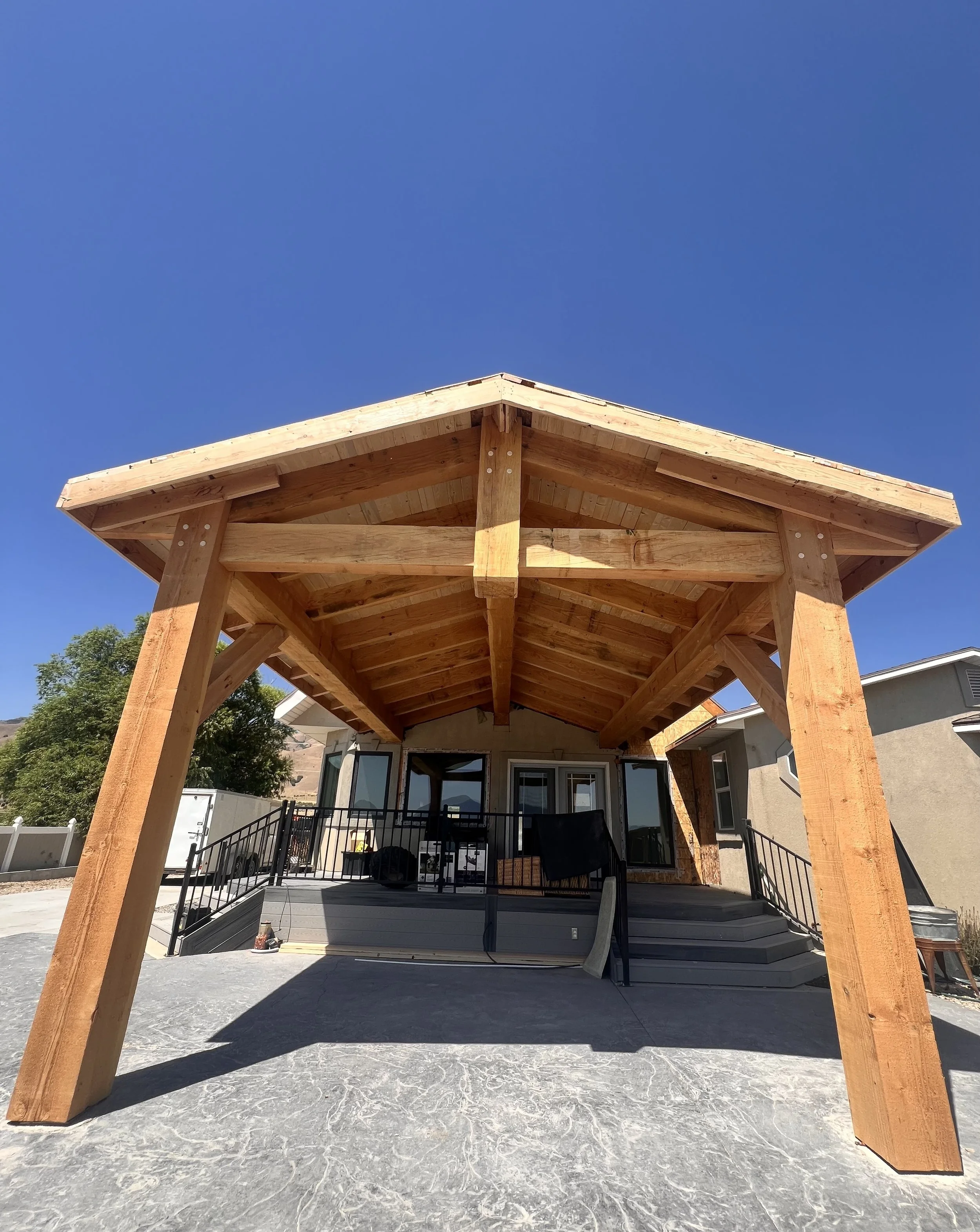Under construction wooden porch roof with support beams over a house porch, with stairs, a black railing, and scattered furniture under construction, on a concrete patio, under a clear blue sky.