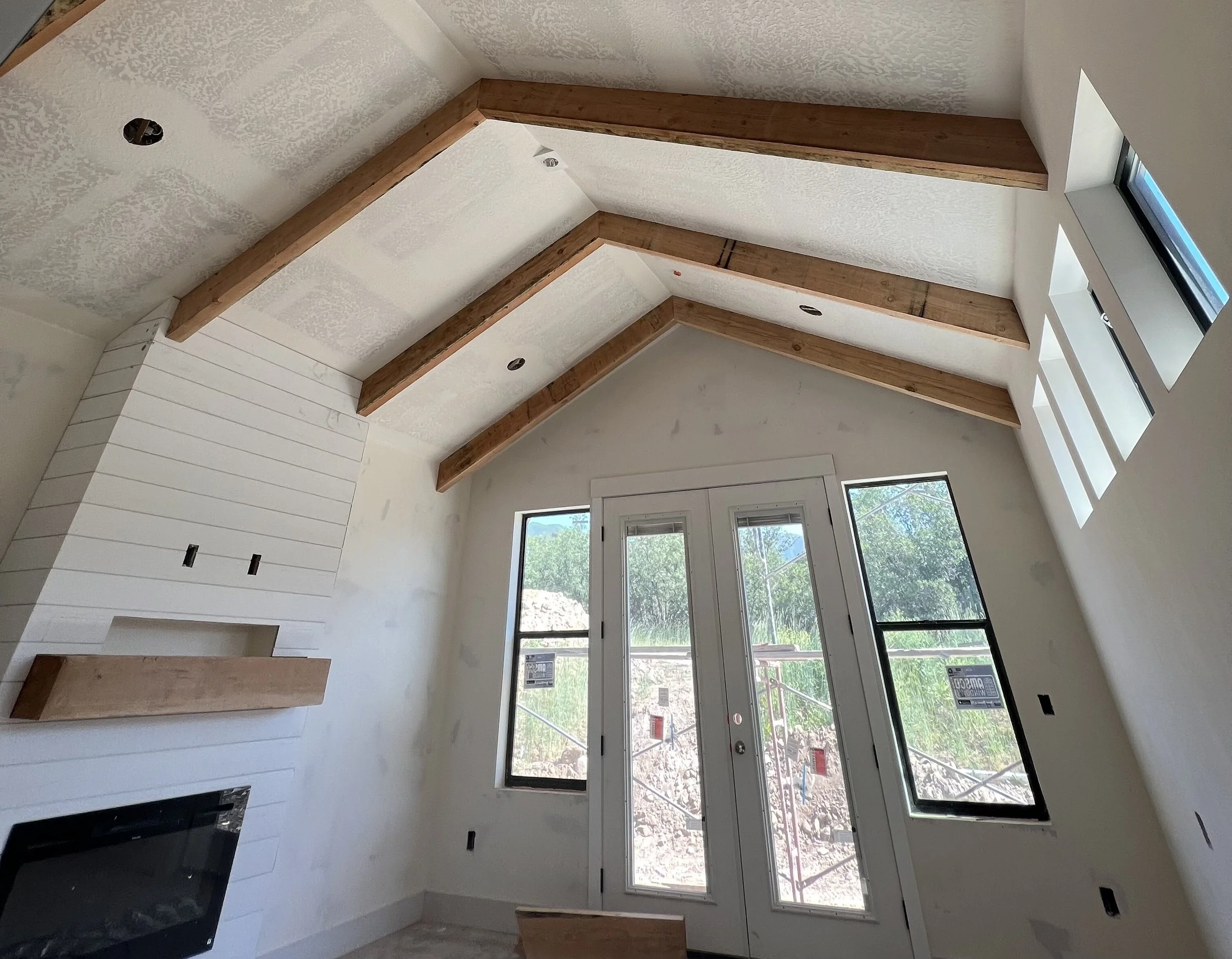 Interior of a room under construction with a vaulted ceiling featuring exposed wooden beams, large windows, and a door leading outside.