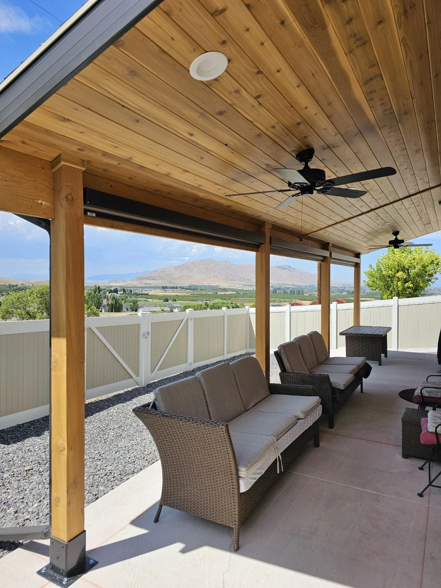 An outdoor covered patio with wicker and cushioned seating, ceiling fans, and a wooden ceiling; overlooking a scenic landscape with mountains, trees, and a white fence.
