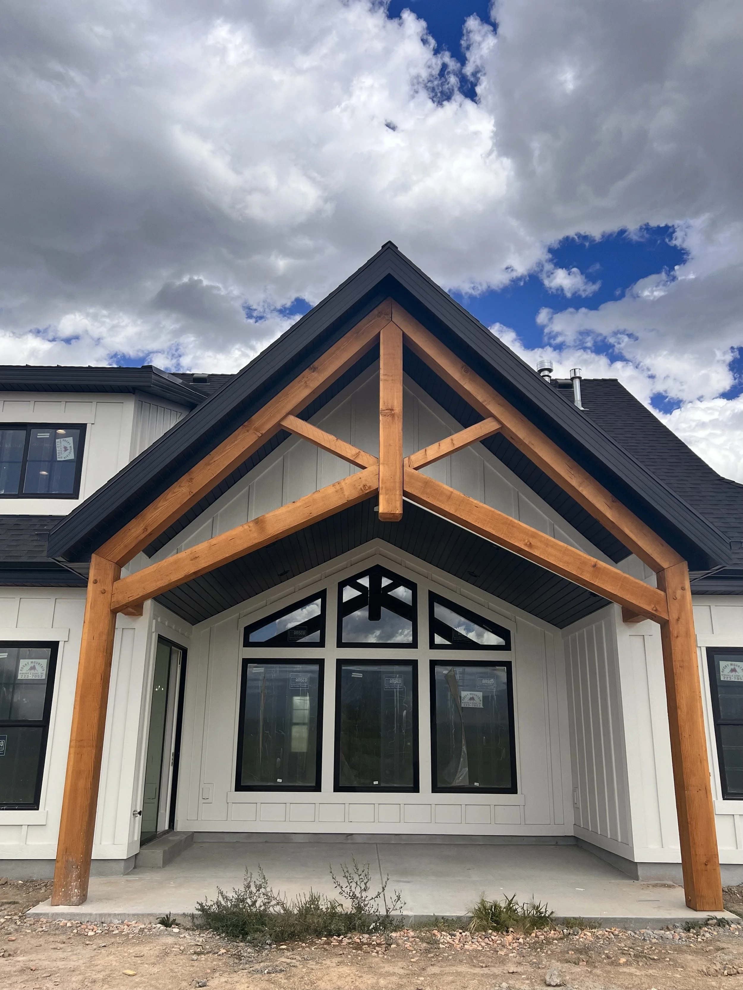 Front view of a house under construction with a large wooden support structure on the porch, white exterior walls, and a peaked roof with a cloudy sky in the background.