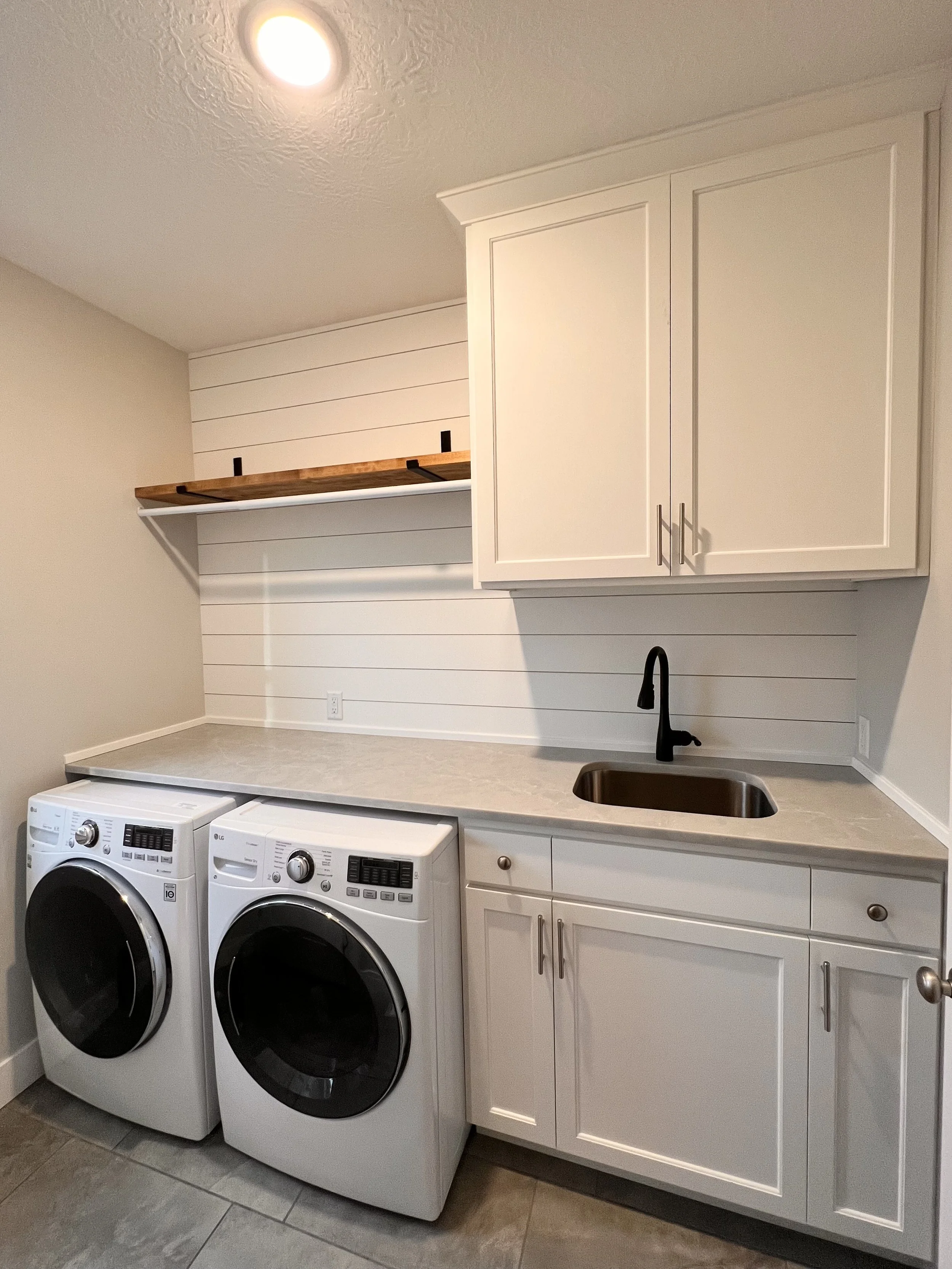 Laundry room with washing machine and dryer, white cabinets, black faucet, countertop, and open wooden shelf above.