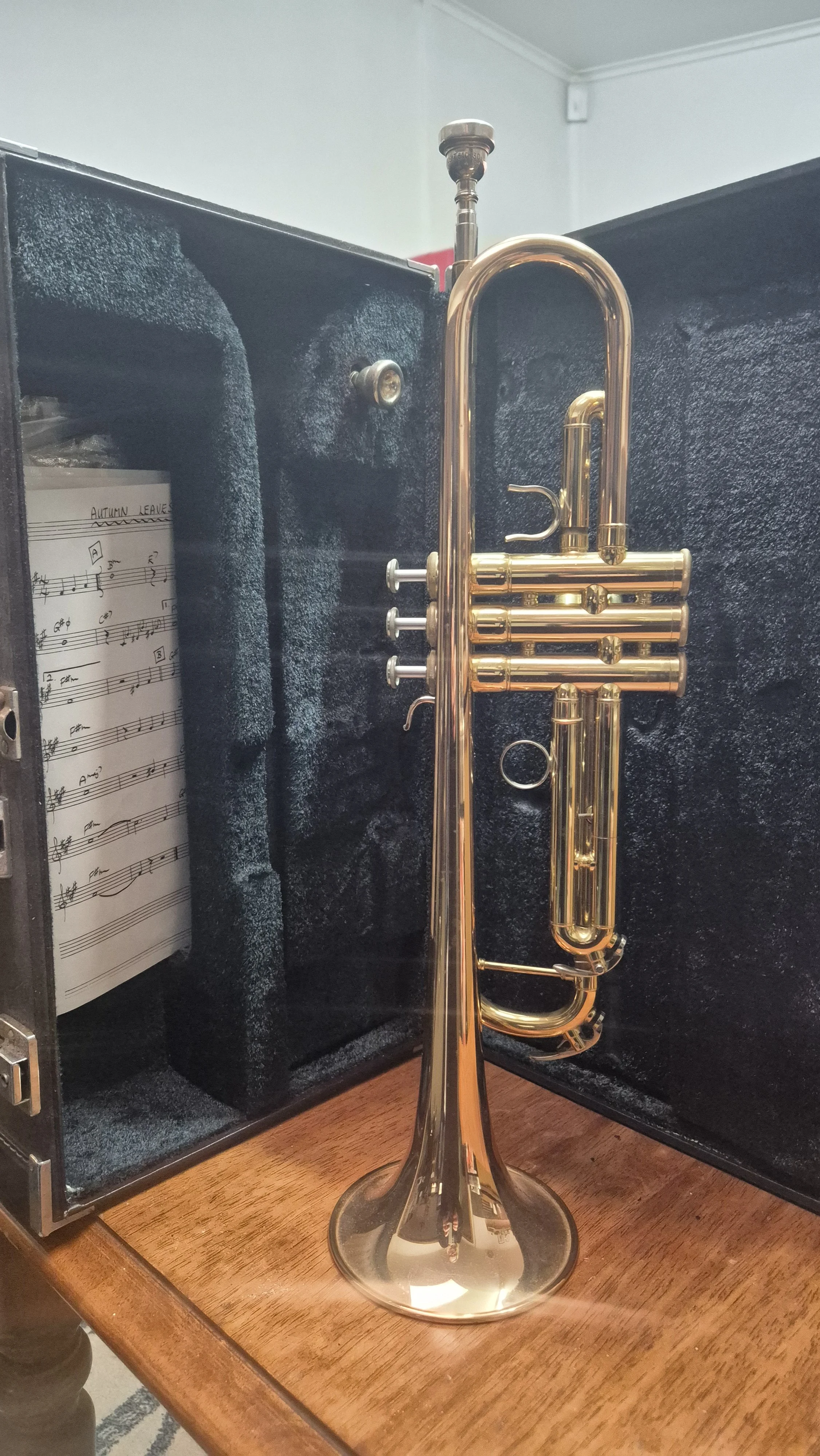 Brass trumpet standing on a wooden surface, open storage case with sheet music inside, and a small red object in the background.