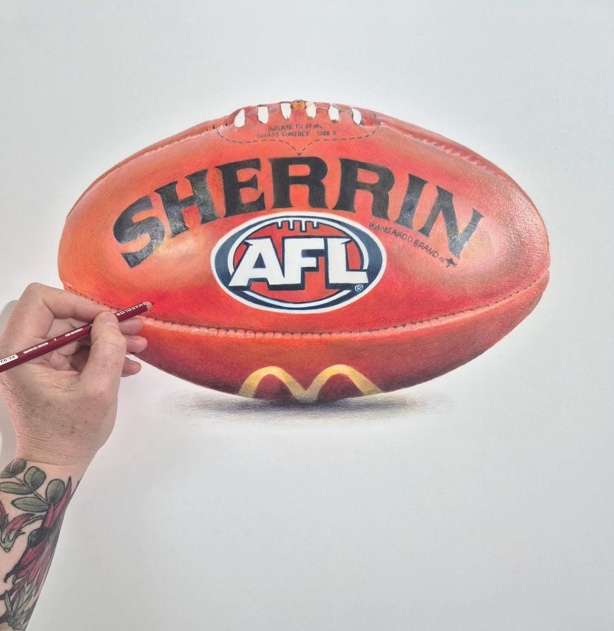 A person's hand holding a red leather football with black and white lettering. The football has a label indicating to 'Insert to amp' and 'Laces Surface Size 1'. The background is a plain white wall.