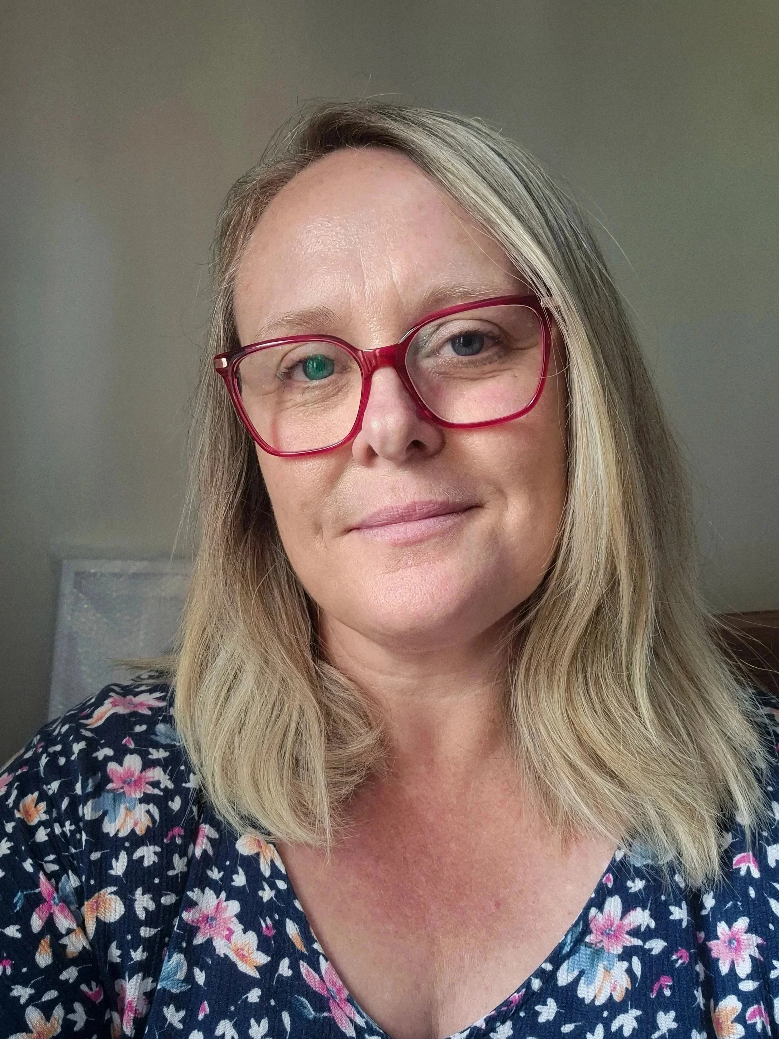 A woman with blonde hair wearing red glasses and a dark floral top, smiling slightly, in a room with a neutral wall.