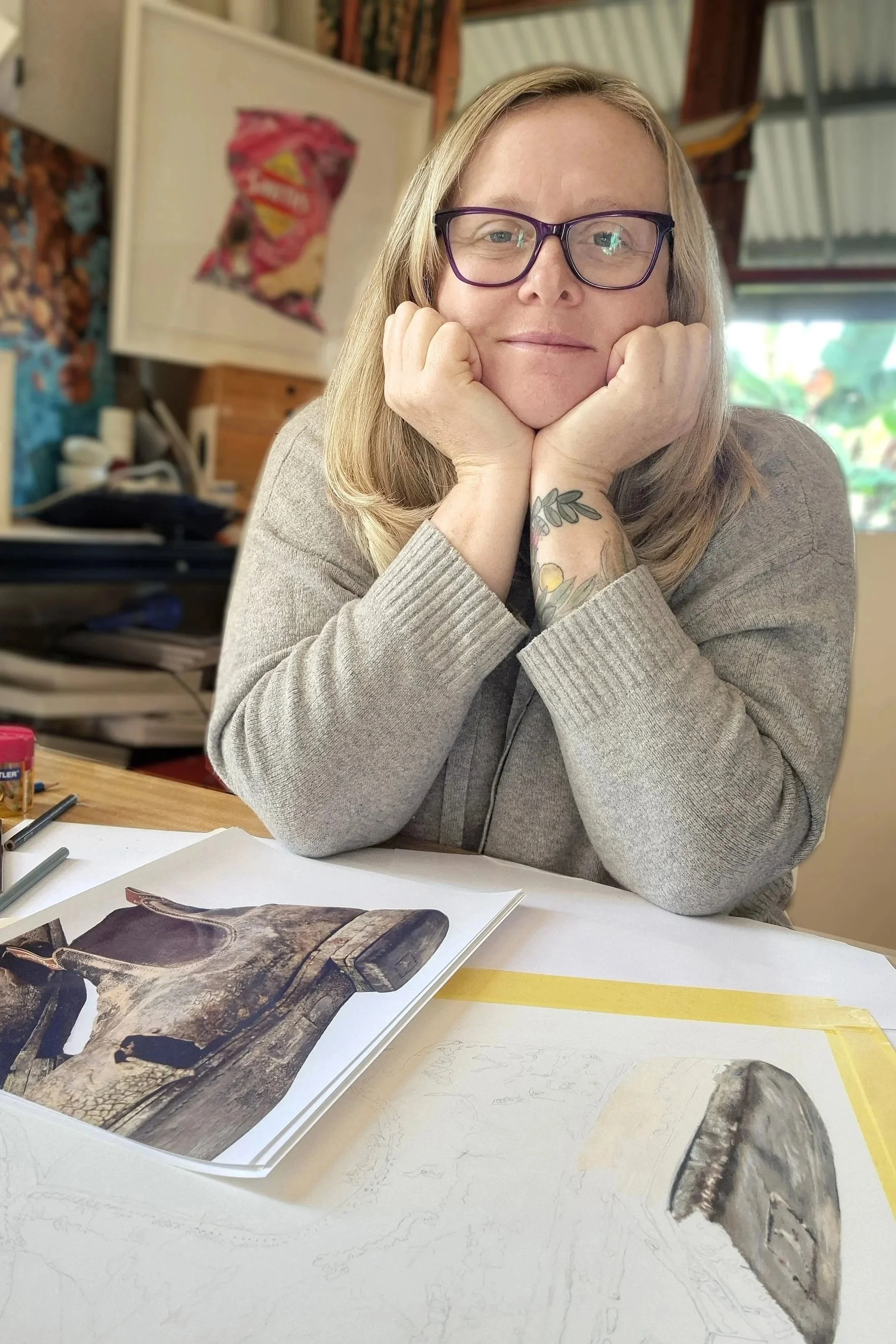 A woman with glasses and blonde hair, sitting at a table with a sketchbook and reference photo of a crocodile. She rests her face in her hands and looks at the camera.