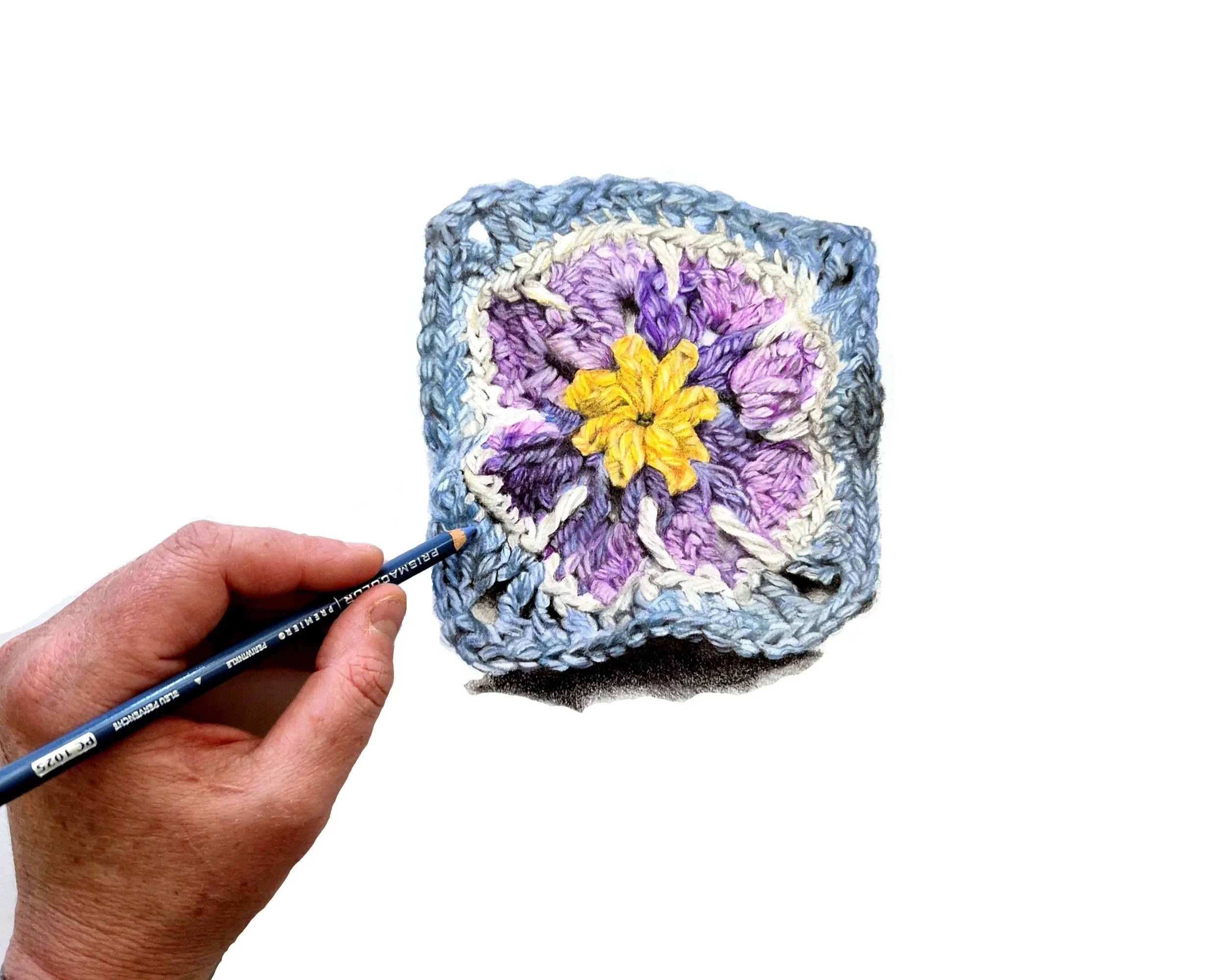 Close-up of a hand holding a colored pencil near a small embroidered piece with purple and yellow floral design on a white background.