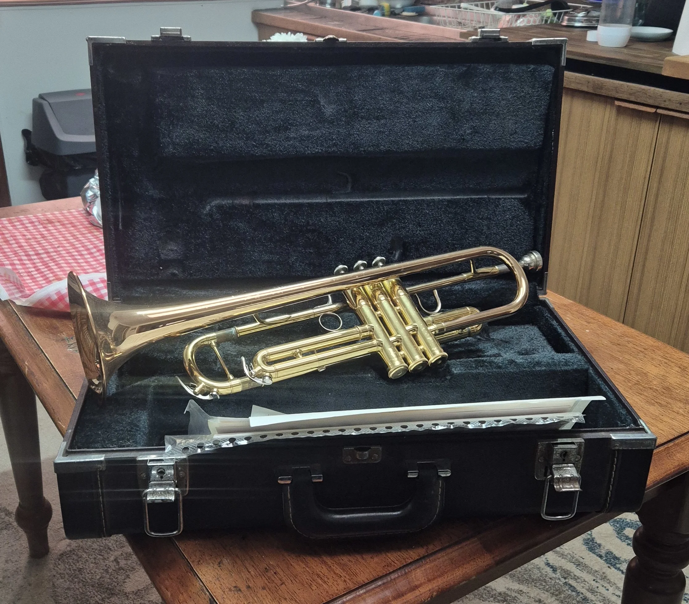 A brass trumpet placed on a wooden table inside a black hard case with a soft interior lining. The case is open, displaying the trumpet. The background includes kitchen items on a counter and a chair near the table.