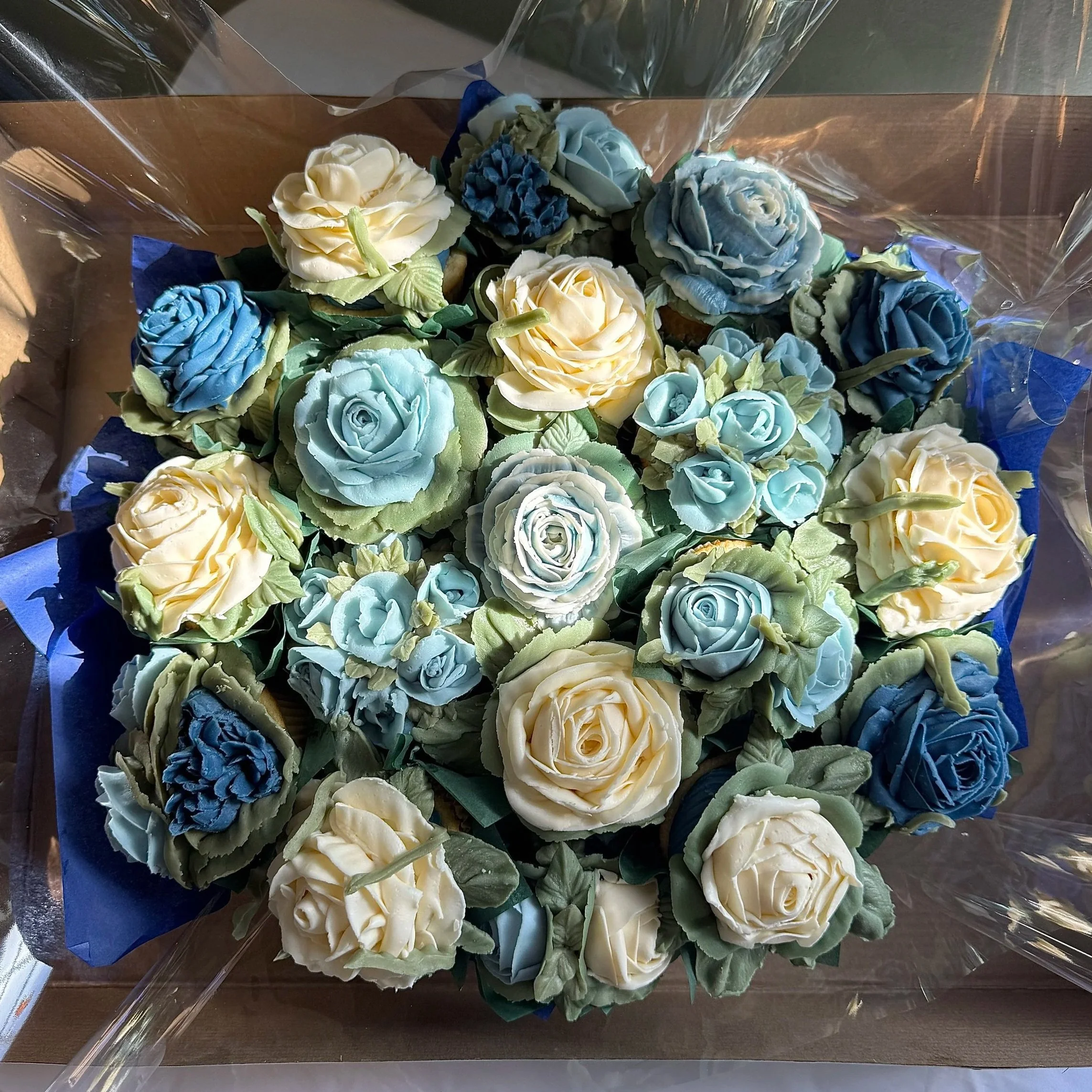 A bouquet of floral cupcakes in shades of dark blue, light blue cream and cream, wrapped in clear cellophane with a blue tissue paper backing.