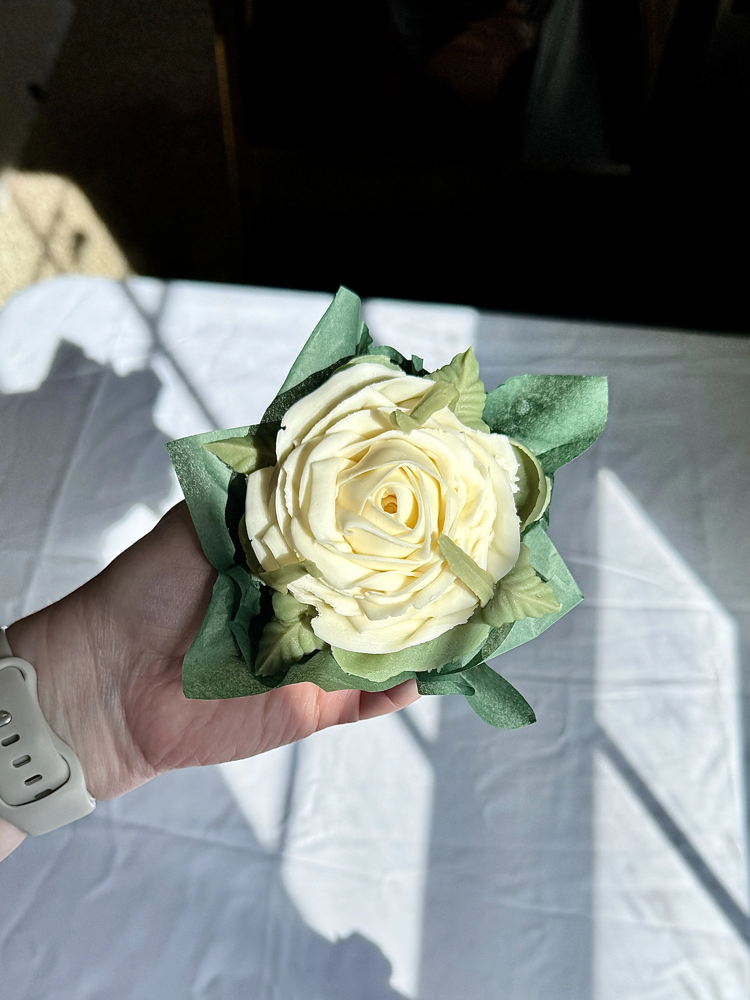 A baker's hand holding a floral cupcake featuring a cream colored rose and green leafing.