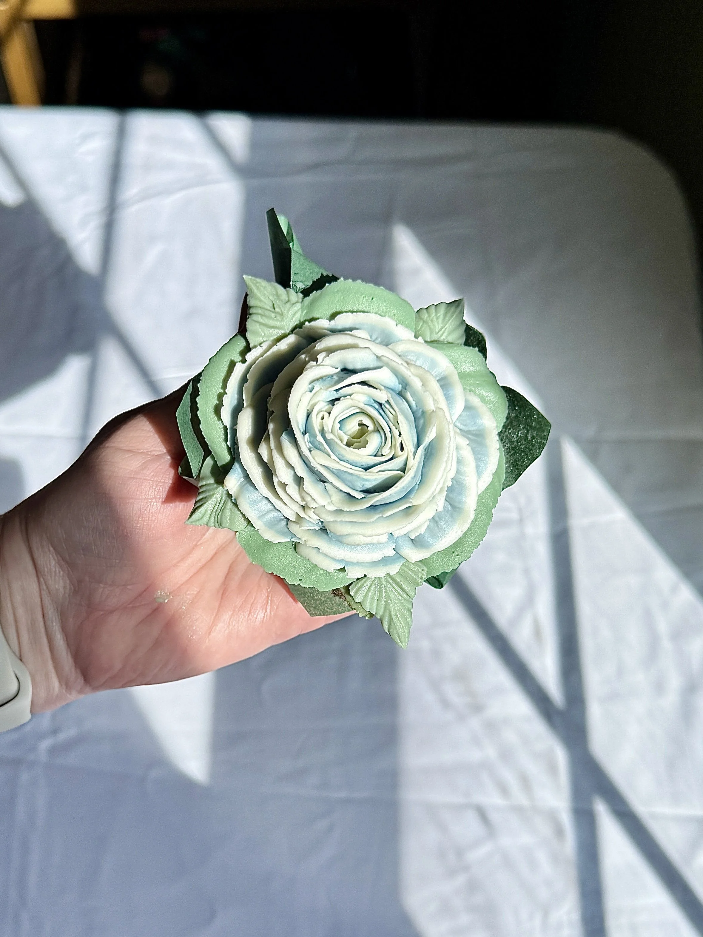 A baker's hand holding a floral cupcake with light blue and white petals and green leaves.
