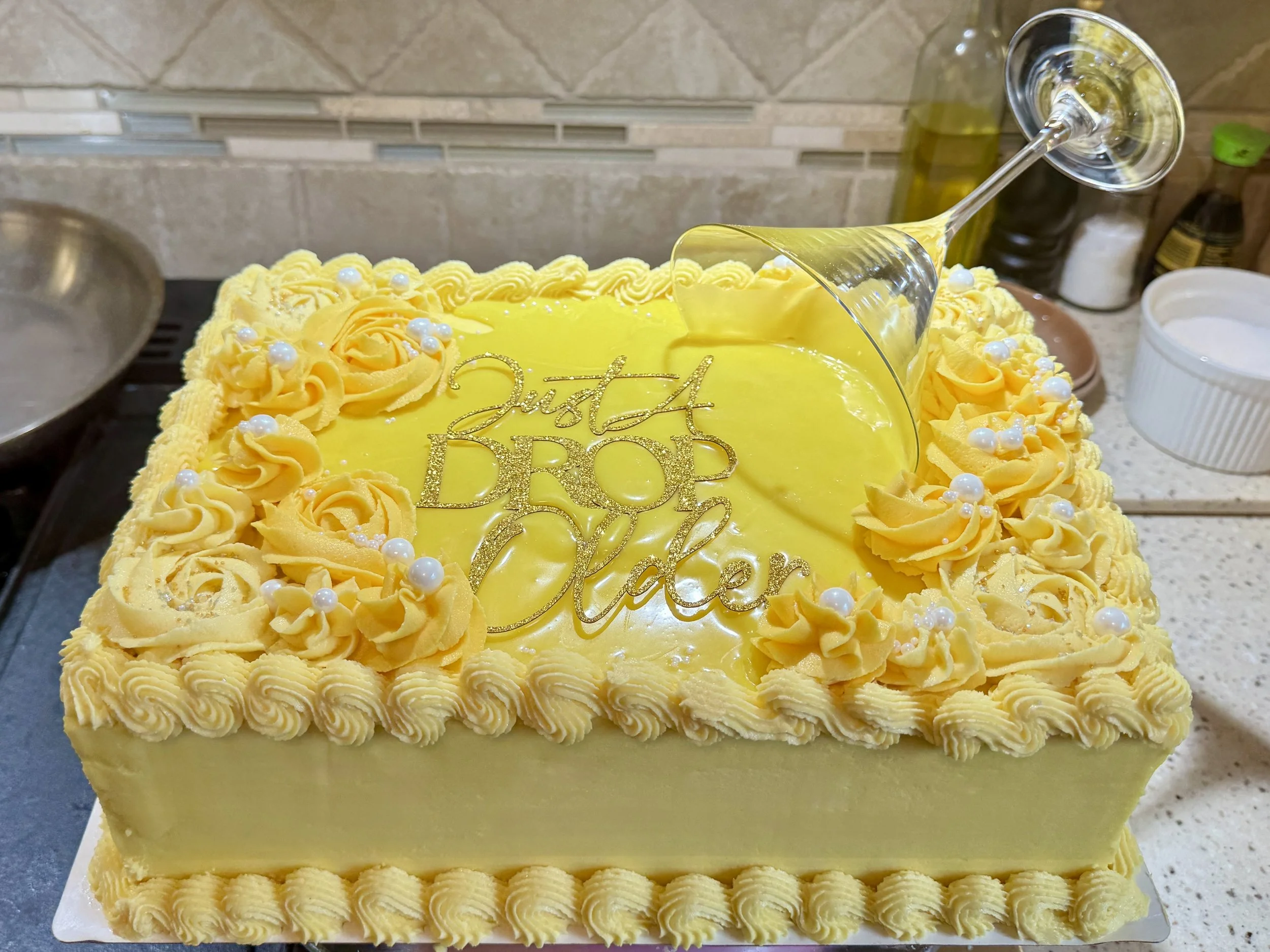 A rectangular lemon yellow birthday cake with a martini glass on top spilling yellow colored white chocolate ganache on to the top of the cake. There is a gold topper that says 'Just a Drop Older.'