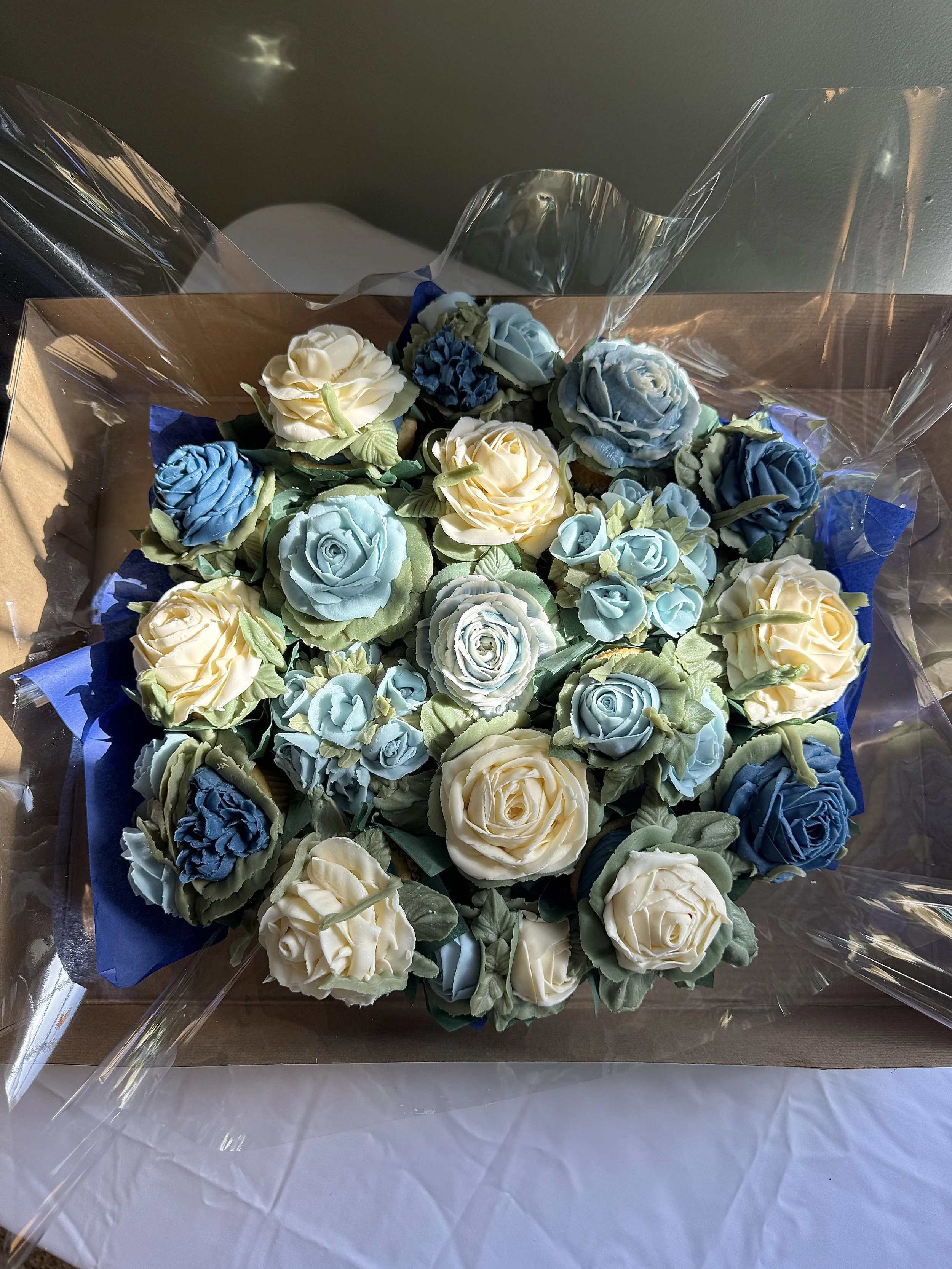 A bouquet of floral cupcakes in shades of dark blue, light blue cream and cream, wrapped in clear cellophane with a blue tissue paper backing.