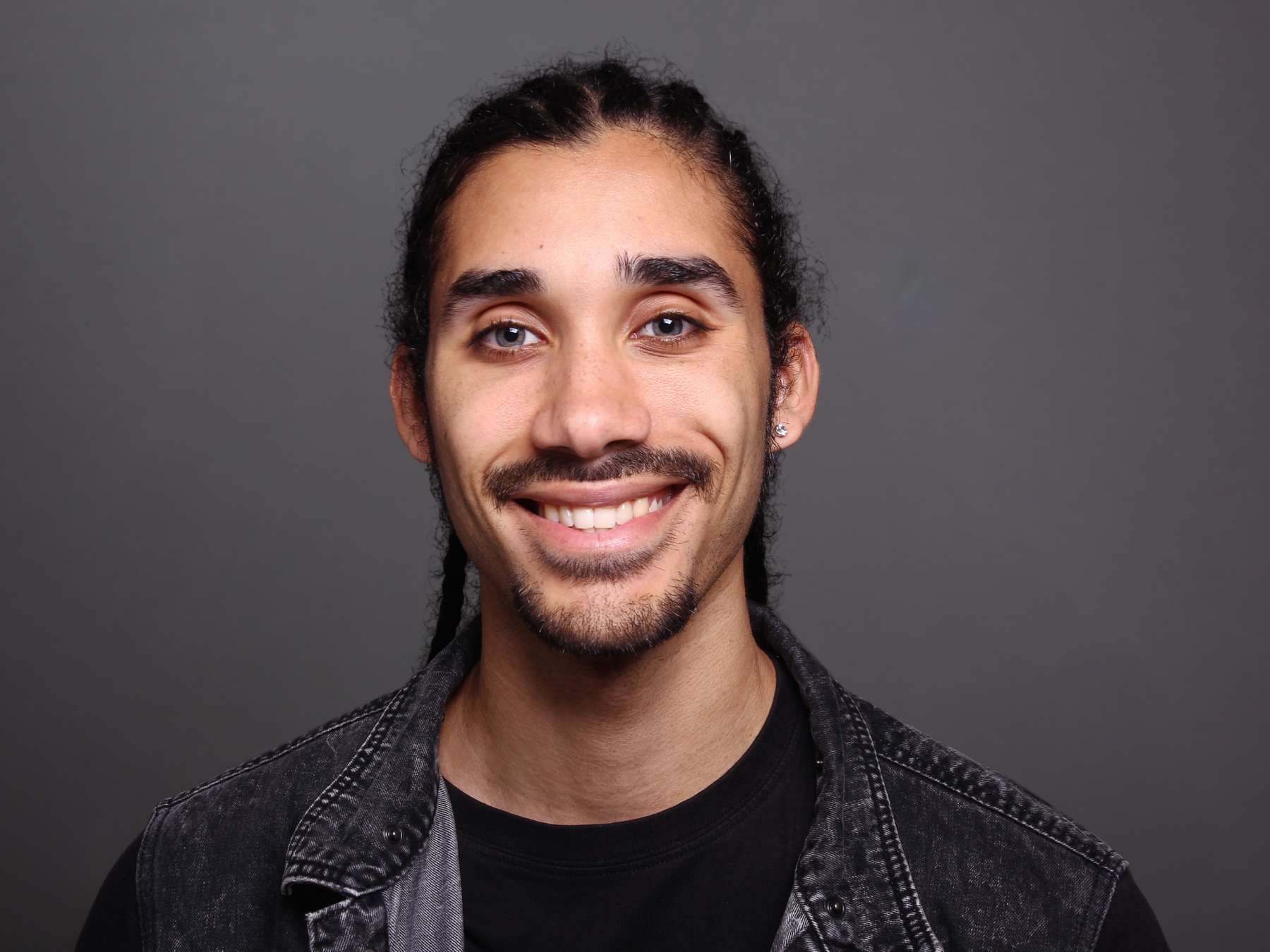 Portrait of a young man with dark curly hair, blue eyes, a goatee, and earrings, wearing a black t-shirt and a black denim jacket, smiling against a plain gray background.