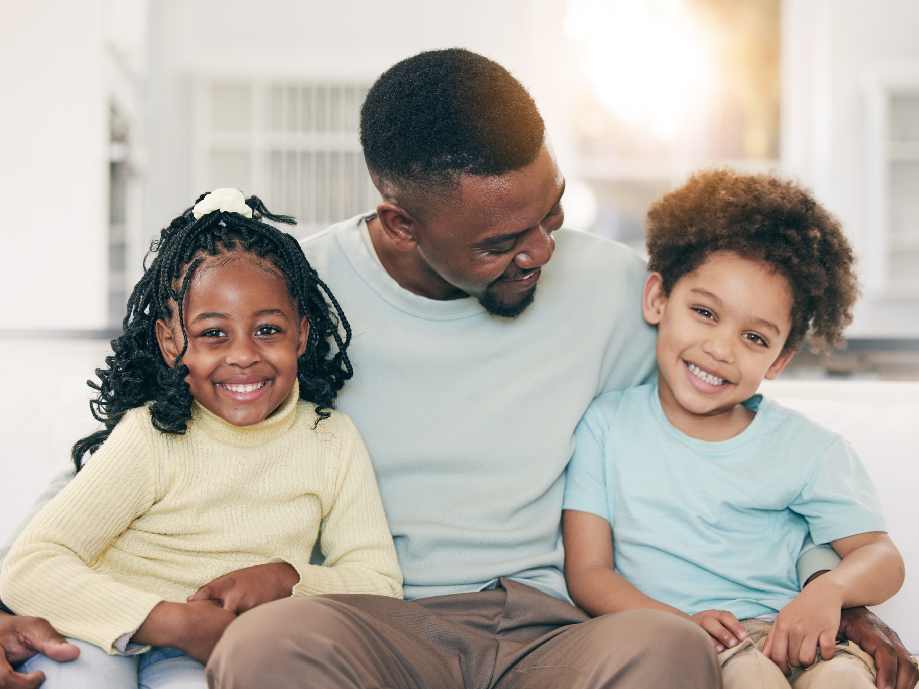 A man with two children, a girl and a boy, sitting closely together on a couch, smiling and looking happy in a bright room.