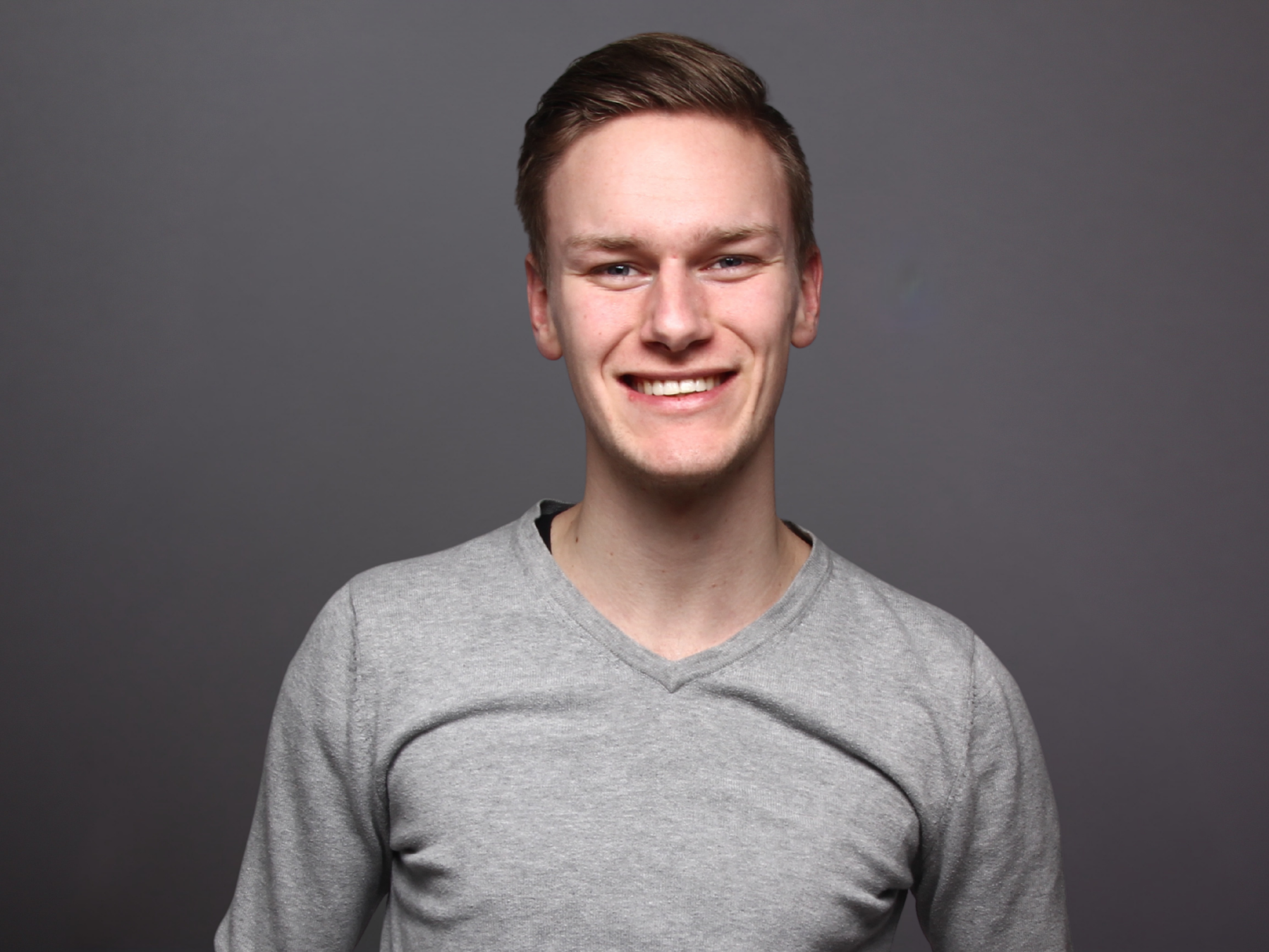 Portrait of a young man with short brown hair, smiling, wearing a light gray V-neck shirt, against a plain gray background.