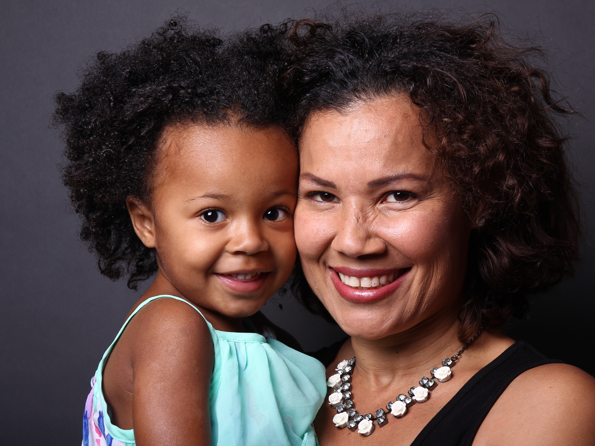A smiling woman holding a young girl with curly hair in front of a dark background.