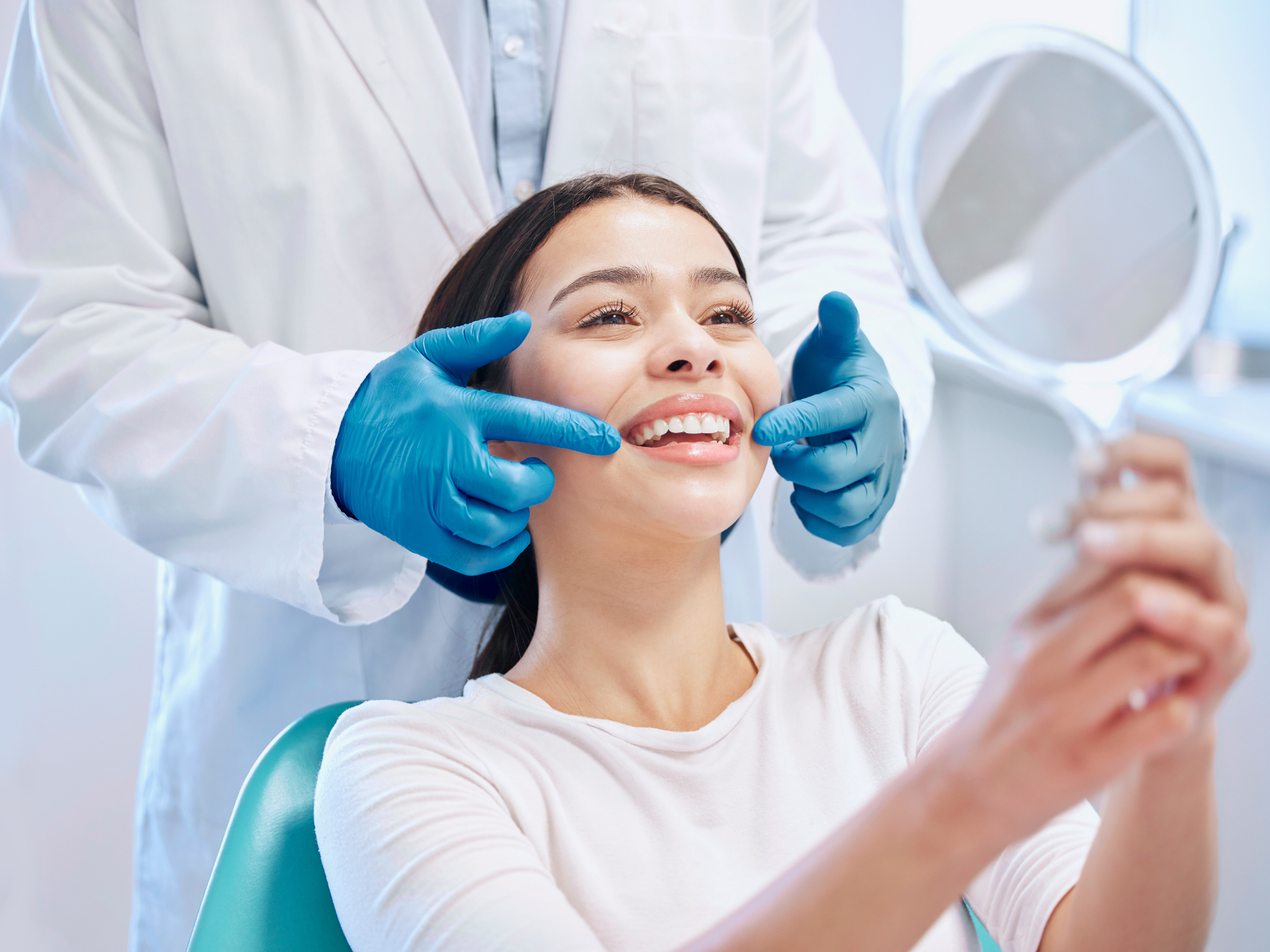 A woman sitting in a dental chair taking a selfie while a dentist examines her teeth and smiles.