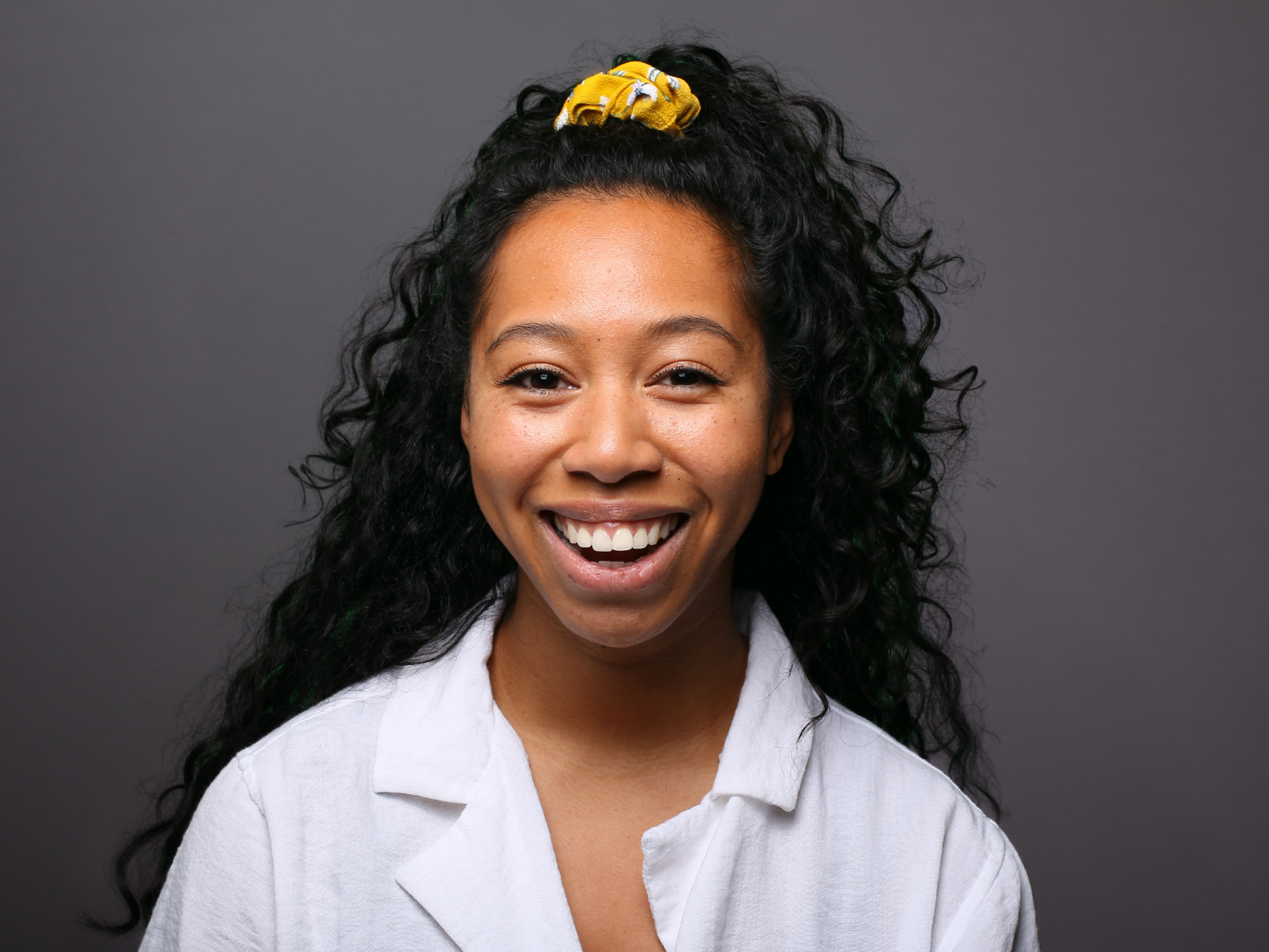 A young woman with dark curly hair, smiling with her mouth open, wearing a white shirt and a yellow scrunchie on top of her head, against a gray background.