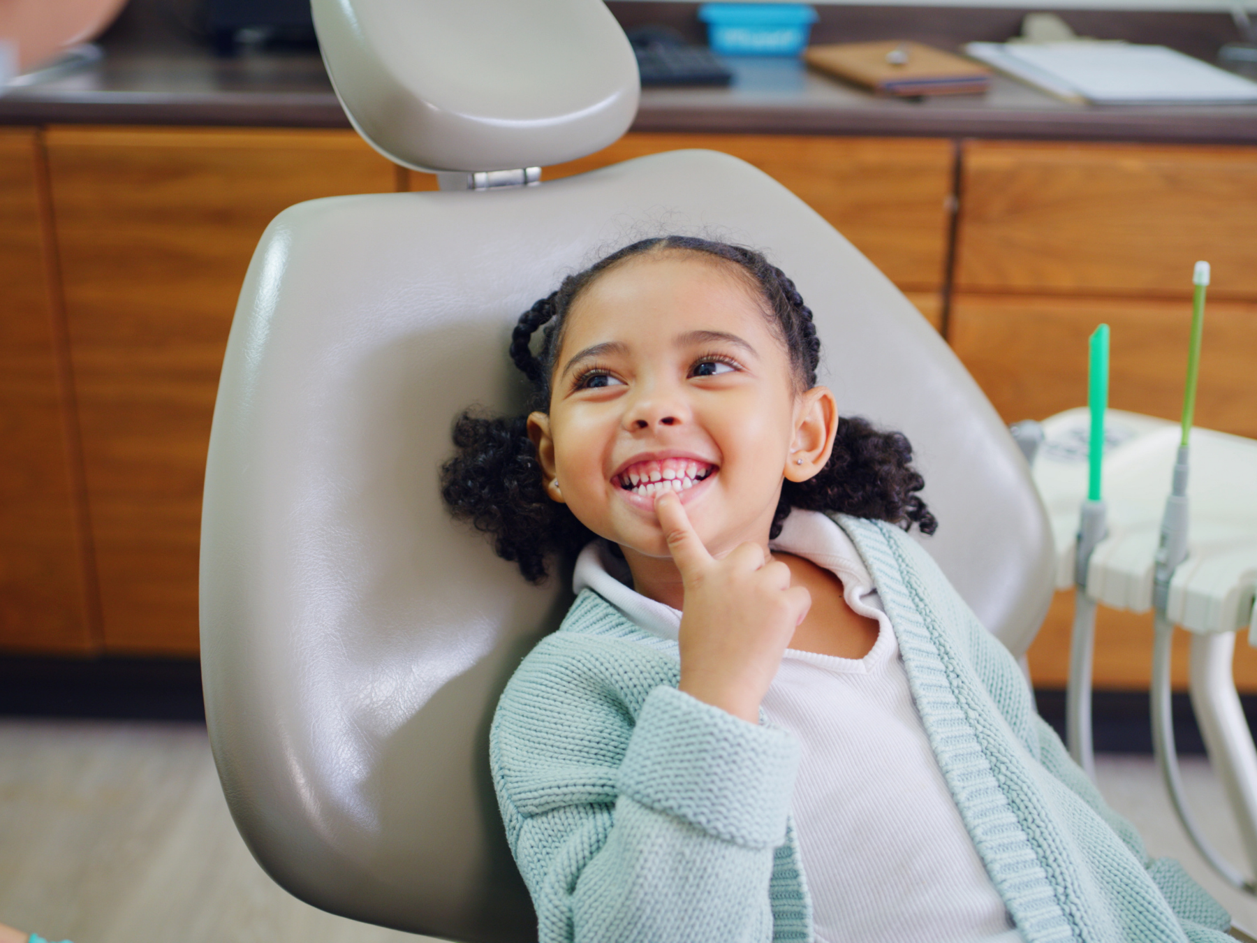 Young girl with curly hair smiling and touching her chin, seated in a dental chair at a dentist's office, with dental tools nearby.